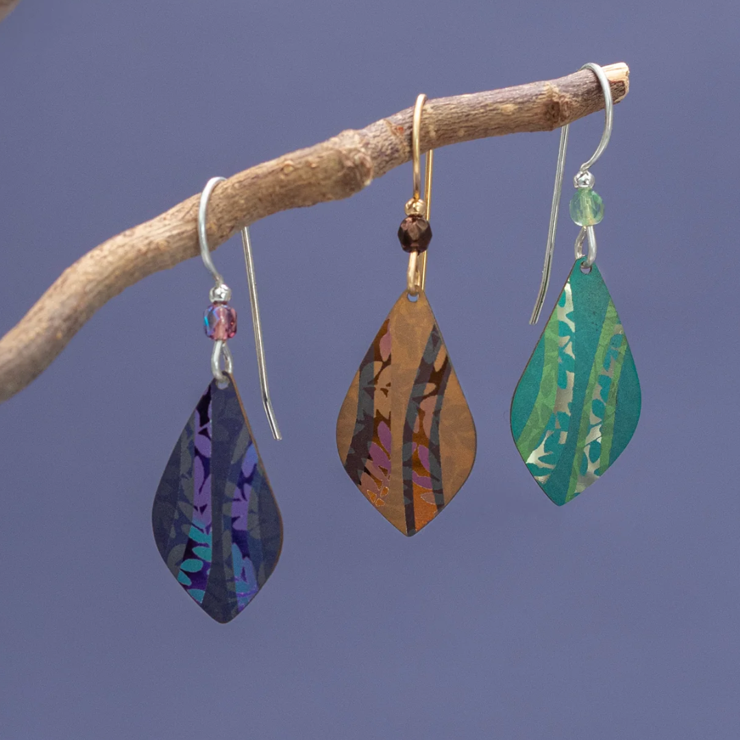 Three vibrant Holly Yashi Riverwind Earrings dangle from a branch against a minimalist backdrop. Each leaf-shaped earring showcases a distinct iridescent pattern, featuring shades of blue, brown, and green. These botanical beauties are elegantly embellished with small crystal beads near the hooks.