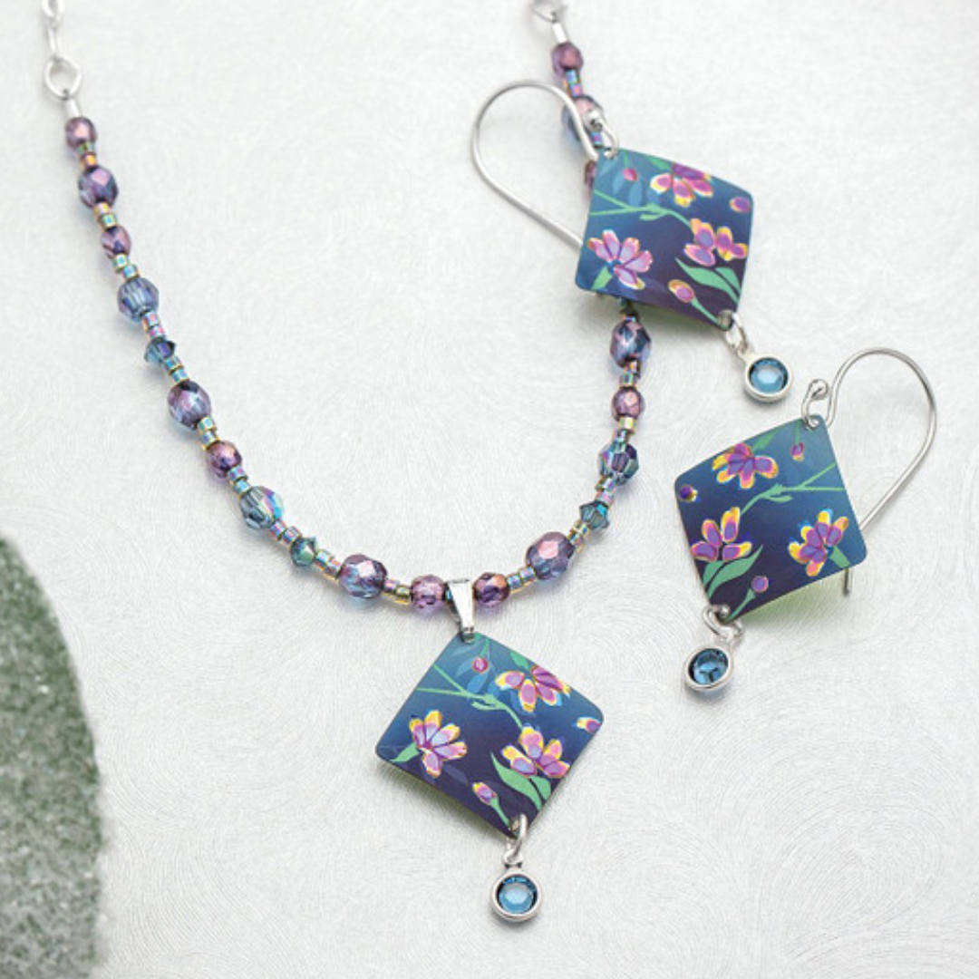 The Holly Yashi Garden Sonnet Beaded Necklace and earring set features square pendants adorned with intricate floral designs. The necklace is accented with purple and blue beads, each pendant graced by a sparkling Swarovski blue gem. The matching earrings perfectly complement the necklace, featuring the same square floral pendants and shimmering blue gems.