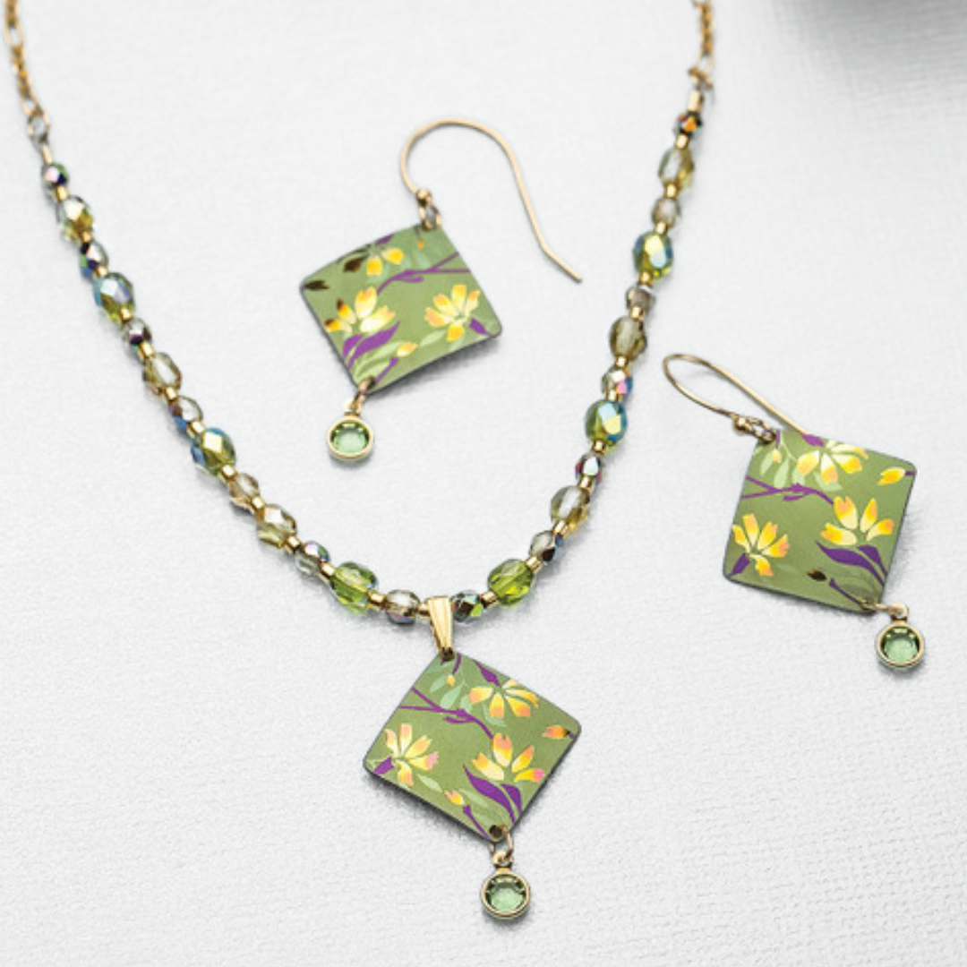 The Holly Yashi Garden Sonnet Beaded Necklace and earring set features a floral design with a touch of Swarovski sparkle. The earrings and pendant showcase a square, green background adorned with yellow flowers and a dangling green gem, while the beaded necklace is embellished with small, multicolored beads. Displayed on a white surface.