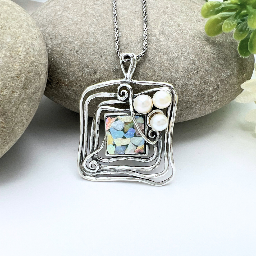 The Square Roman Glass and Pearl Necklace features a silver pendant with an abstract, wavy square design hanging from a handmade sterling setting on a silver chain. The pendant showcases three small white pearls and a mosaic of colorful Roman glass fragments in the center. The necklace is beautifully displayed against a backdrop of smooth grey stones and green foliage.