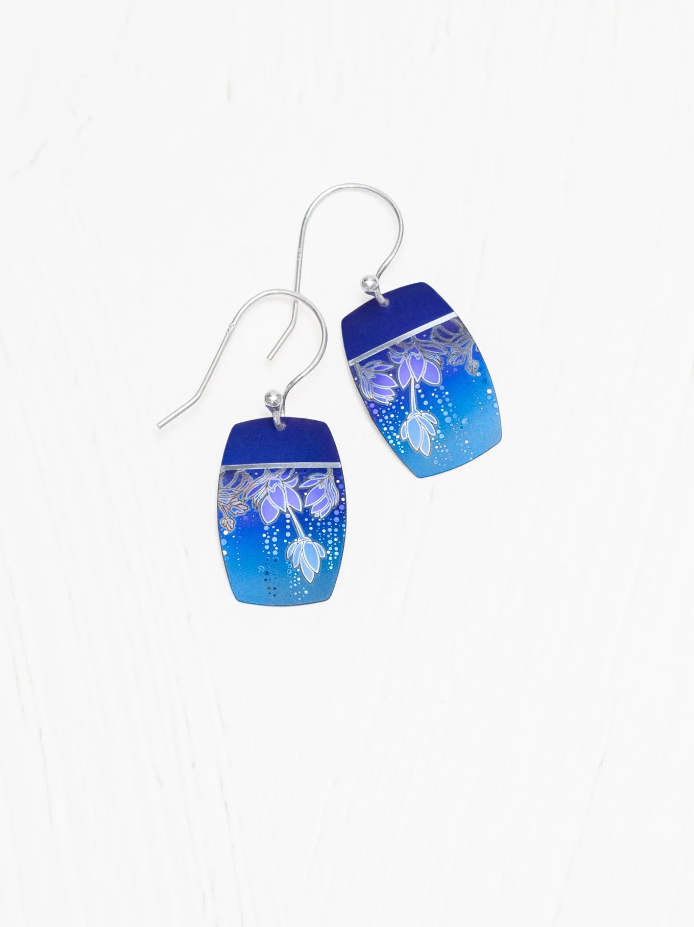 Introducing the Holly Yashi Cari Earrings: A pair of tropical floral earrings with a gradient design that transitions from dark blue at the top to lighter blue at the bottom. Featuring delicate silver floral patterns on a rectangular, slightly curved metal surface with silver hooks, these ocean blues jewelry pieces are truly captivating.