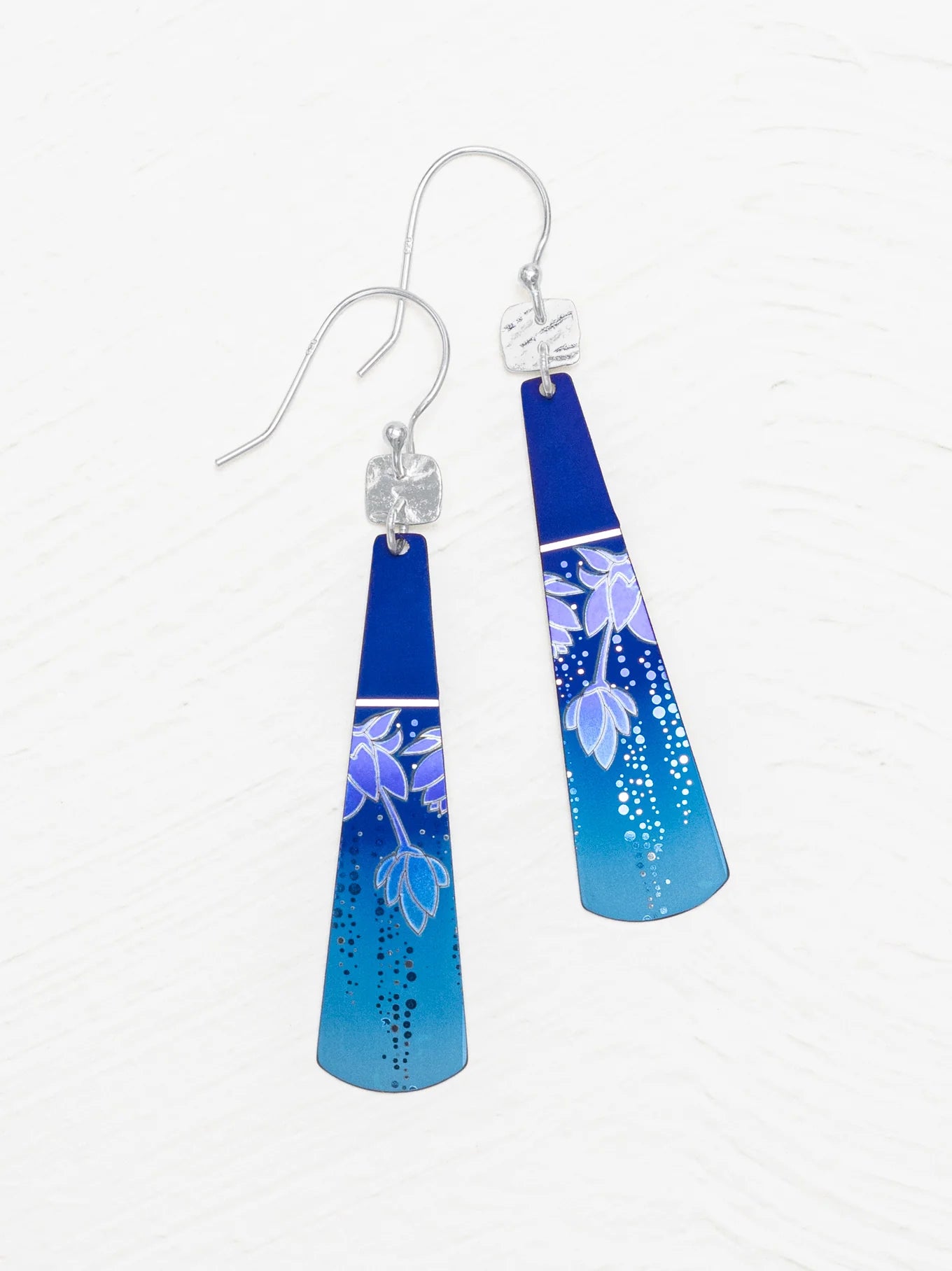Introducing the Holly Yashi Tula Earrings: These exquisite triangular earrings boast a stunning gradient transition from deep blue at the top to light blue at the bottom. Adorning each earring is a delicate floral design in soft light purple and white, enhanced by tiny white dots that accentuate the lower part for a truly romantic look.