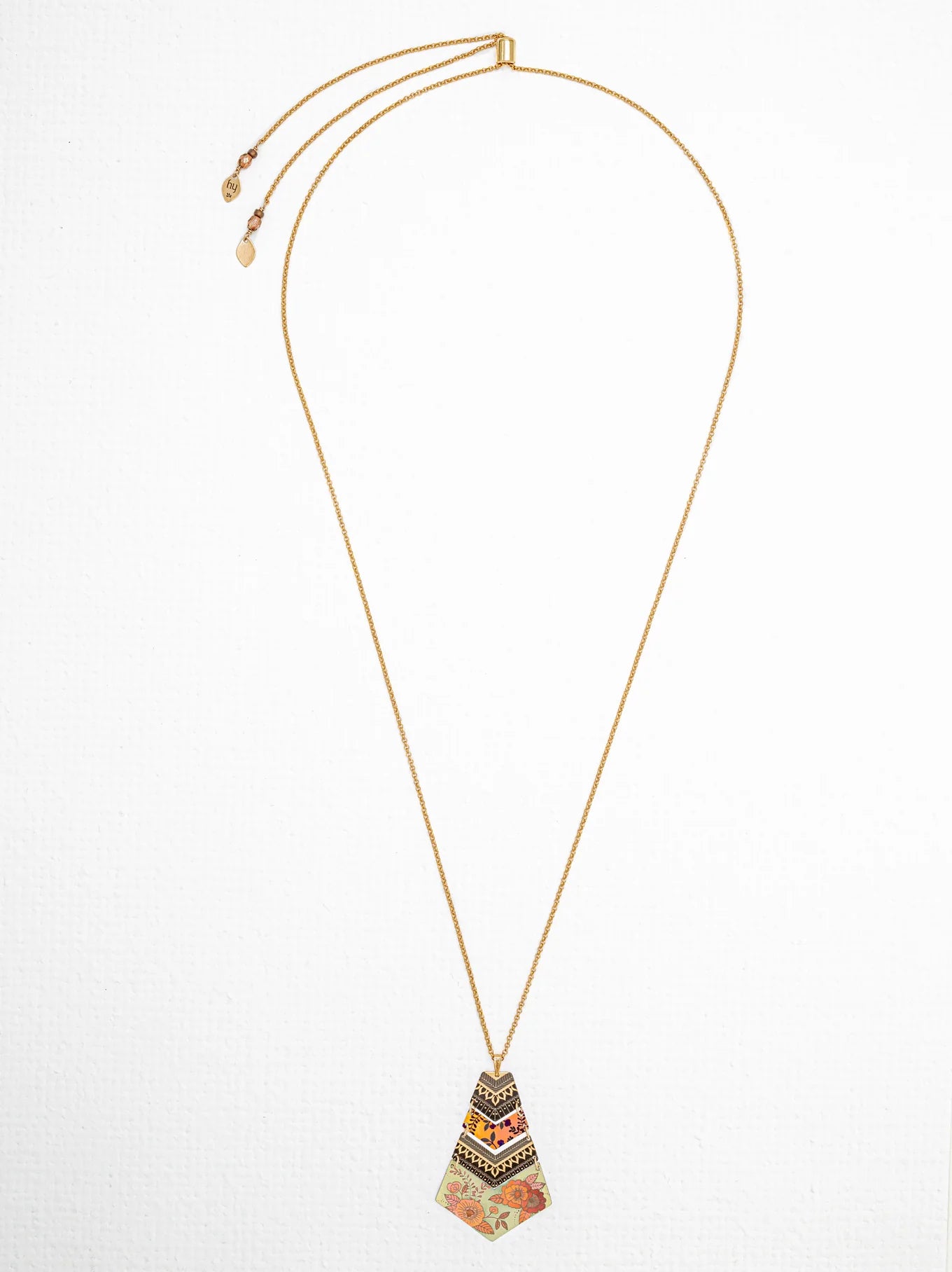The Holly Yashi Avant Garden Necklace is a long Bohemian-inspired piece featuring a uniquely shaped niobium pendant adorned with intricate patterns and vibrant designs in shades of black, gold, and orange. This handcrafted jewelry item includes an adjustable clasp accented with small decorative charms at the ends.