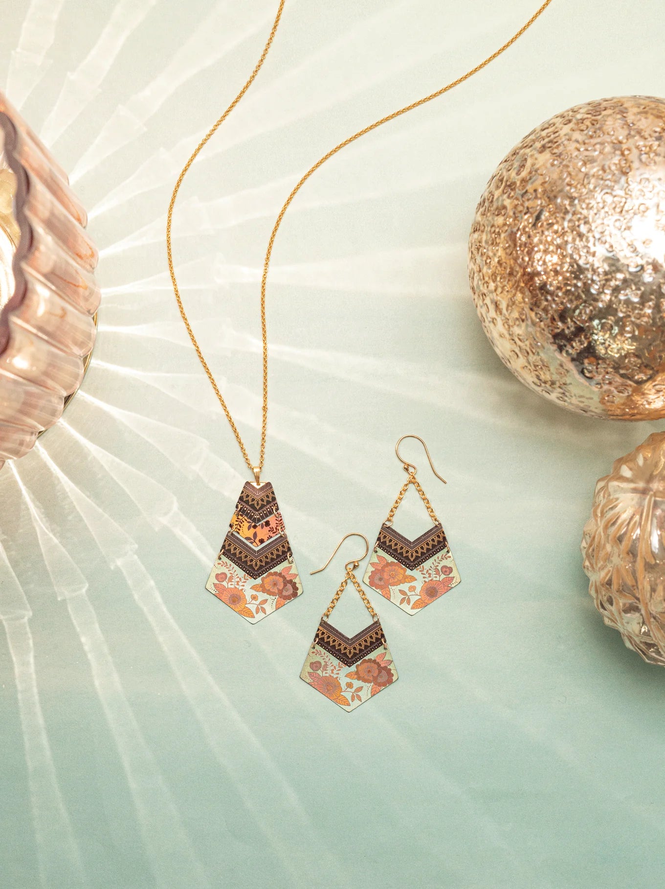 The Holly Yashi Avant Garden Necklace and a pair of earrings with geometric and floral designs are elegantly arranged on a light green surface. The jewelry showcases shades of gold, black, and coral. Nearby, decorative textured glass ornaments in gold tones cast soft light reflections, enhancing the necklace's Bohemian charm.