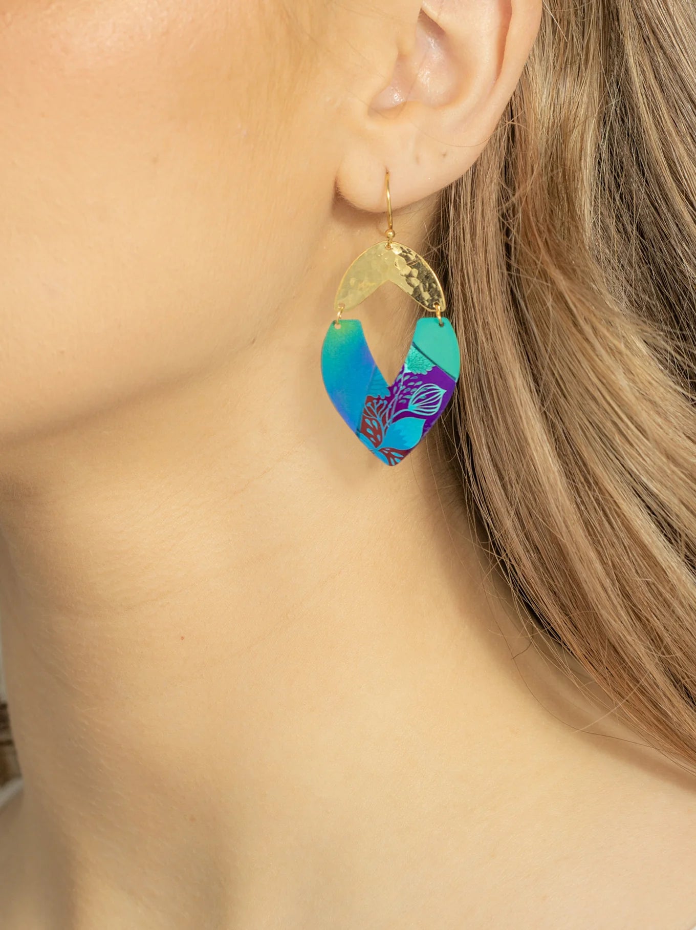 A close-up captures a person’s ear adorned with Holly Yashi Kalea Earrings. These stunning dangle earrings feature a gold piece at the top and a teardrop-shaped section below, showcasing a vibrant blue, purple, and teal leaf pattern reminiscent of island florals. The person’s blonde hair complements their fair skin beautifully.