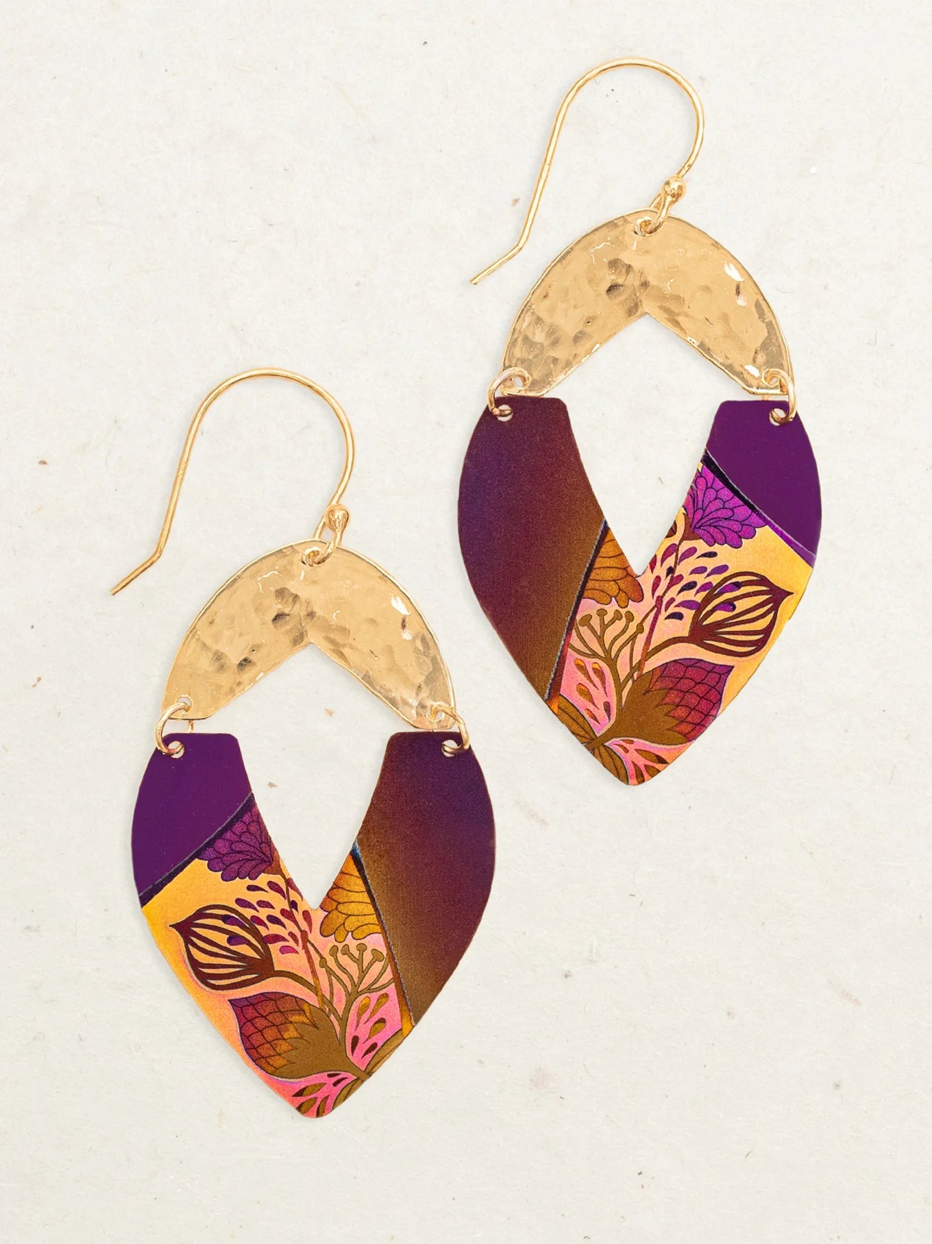 The Holly Yashi Kalea Earrings are showcased against a neutral background. These niobium metal earrings feature an upper gold-toned, textured half-leaf shape, complemented by a larger lower section painted with a gradient of purple and yellow hues adorned with intricate island florals in pink and orange.