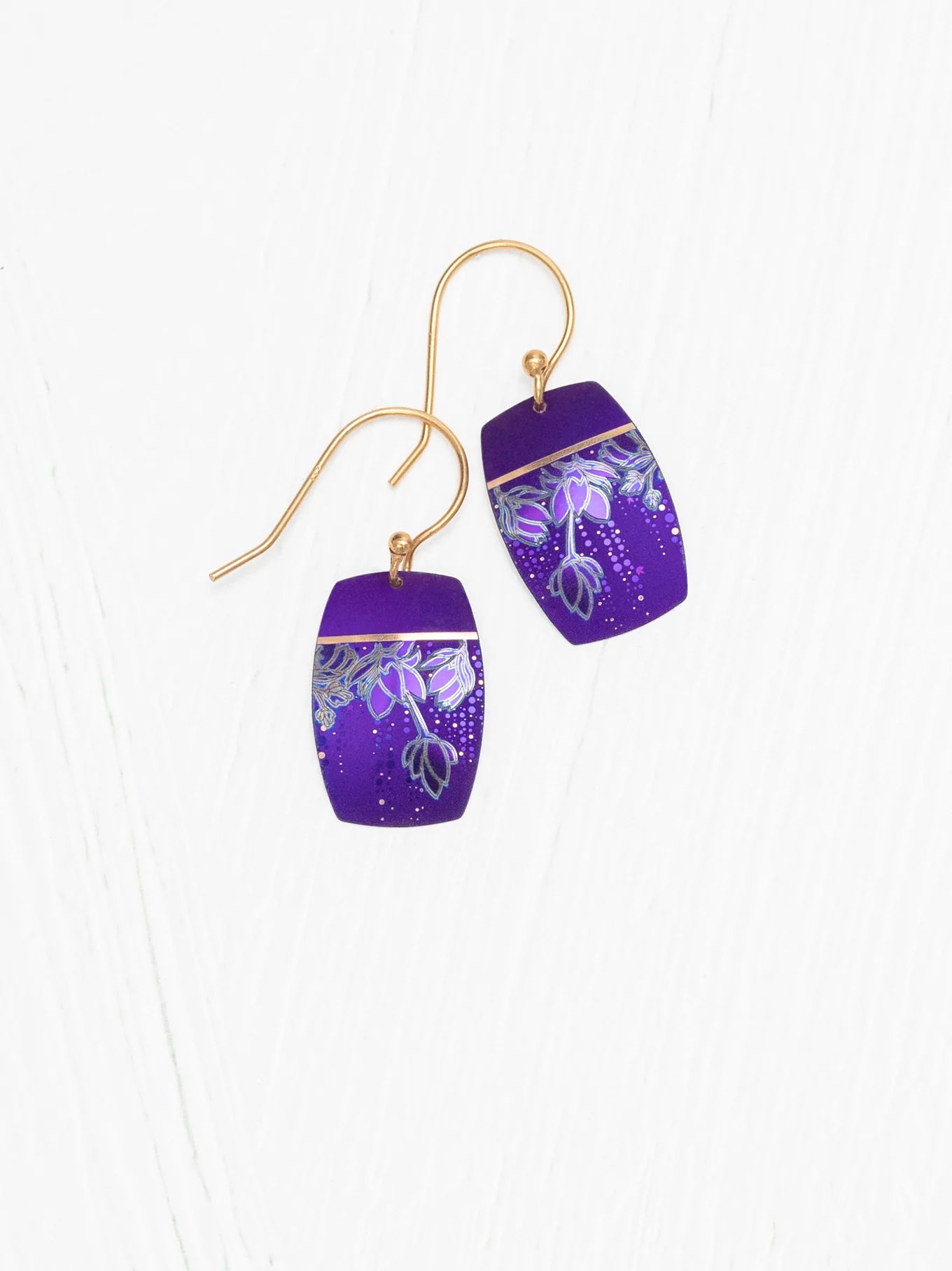 The Holly Yashi Cari Earrings are a pair of purple niobium earrings featuring a tropical floral and leaf design in silver, set against a white background, and fitted with gold hooks.
