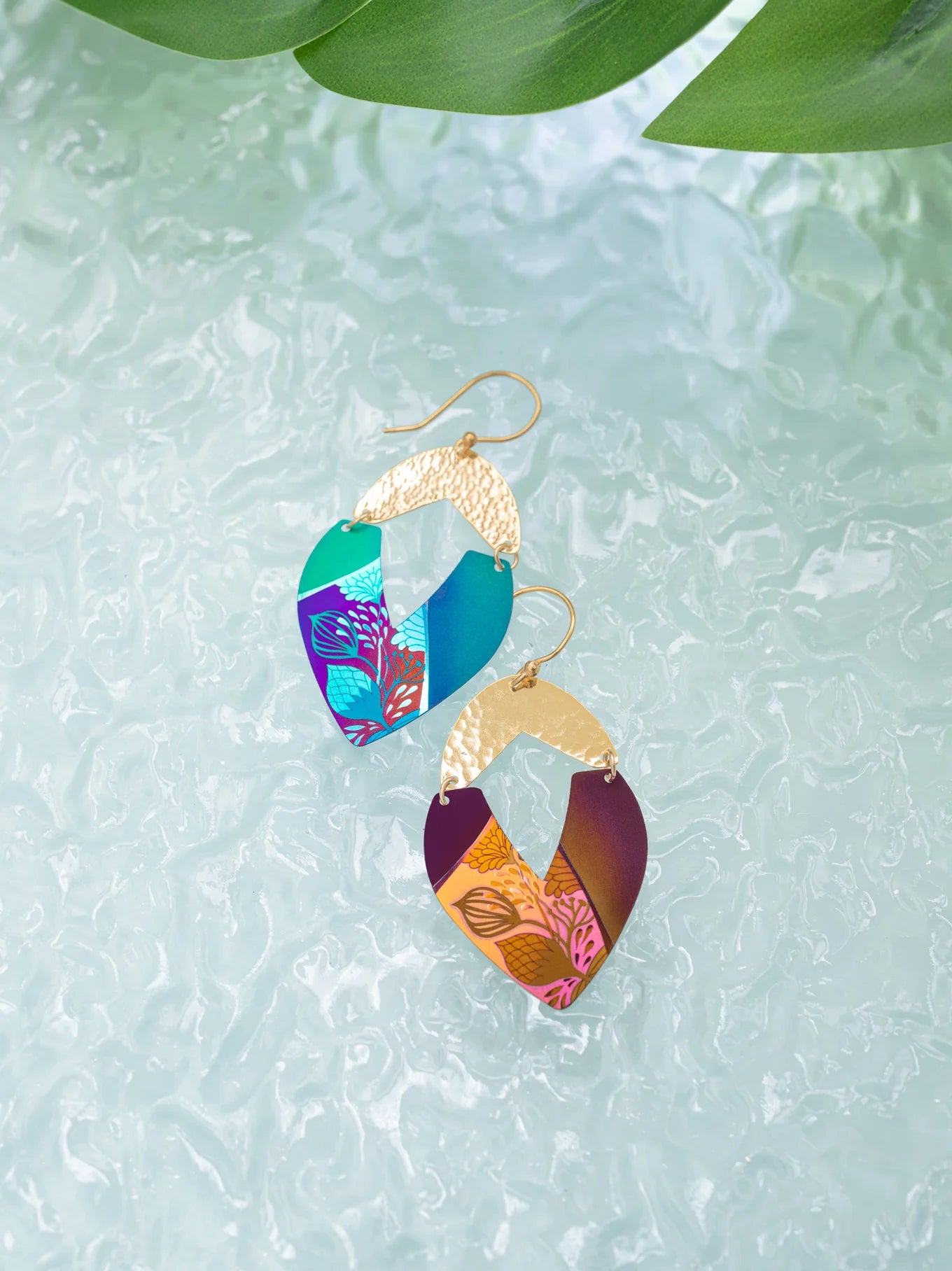 A pair of elegant Holly Yashi Kalea Earrings featuring intricate designs with a hammered gold top section, linking to vibrant blue, purple, and orange hues adorned with detailed leaf-like patterns reminiscent of island florals. The earrings are showcased on a textured, icy background.