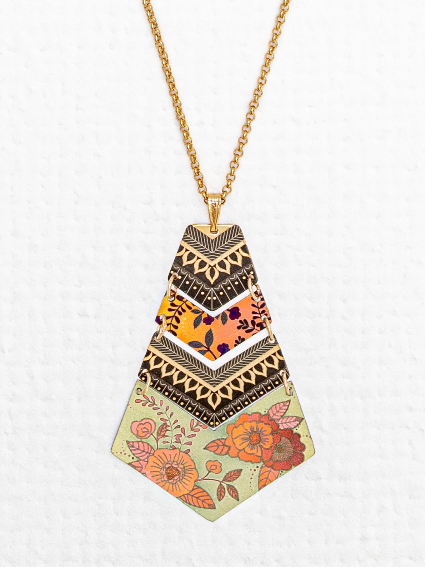 The **Holly Yashi Avant Garden Necklace** is a gold chain with a handcrafted niobium pendant that features an array of intricate patterns. The top section displays black and gold triangular designs, the middle section bursts with vibrant colors and abstract shapes, while the bottom section offers floral patterns on a light green background for a Bohemian touch.
