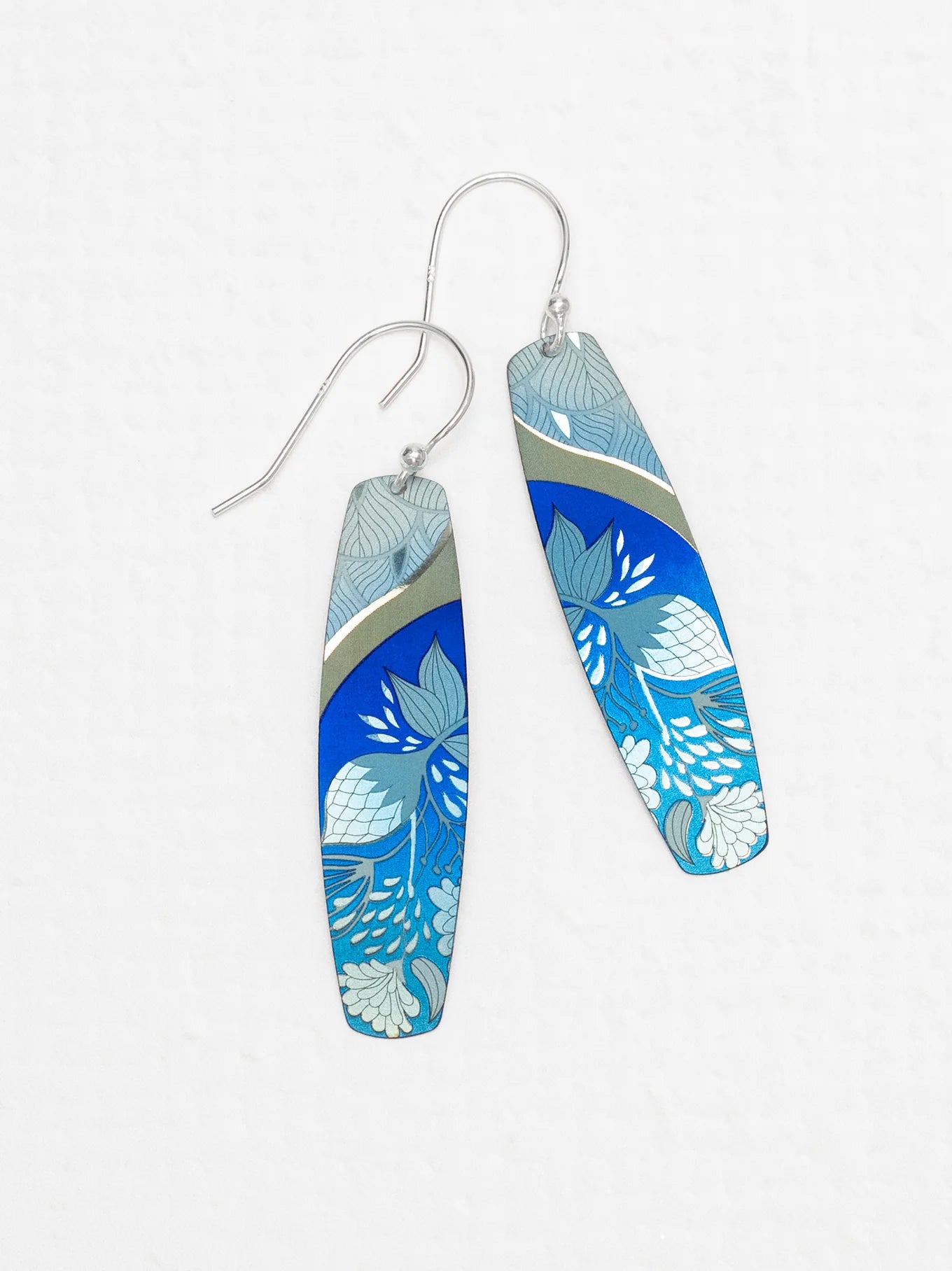 The Holly Yashi Malia Earrings are a stunning pair of elongated teardrop-shaped accessories featuring 18K gold-plated niobium hooks and a vibrant blue, white, and teal tropical floral pattern. Intricate botanical designs with curved silver lines add an elegant touch.
