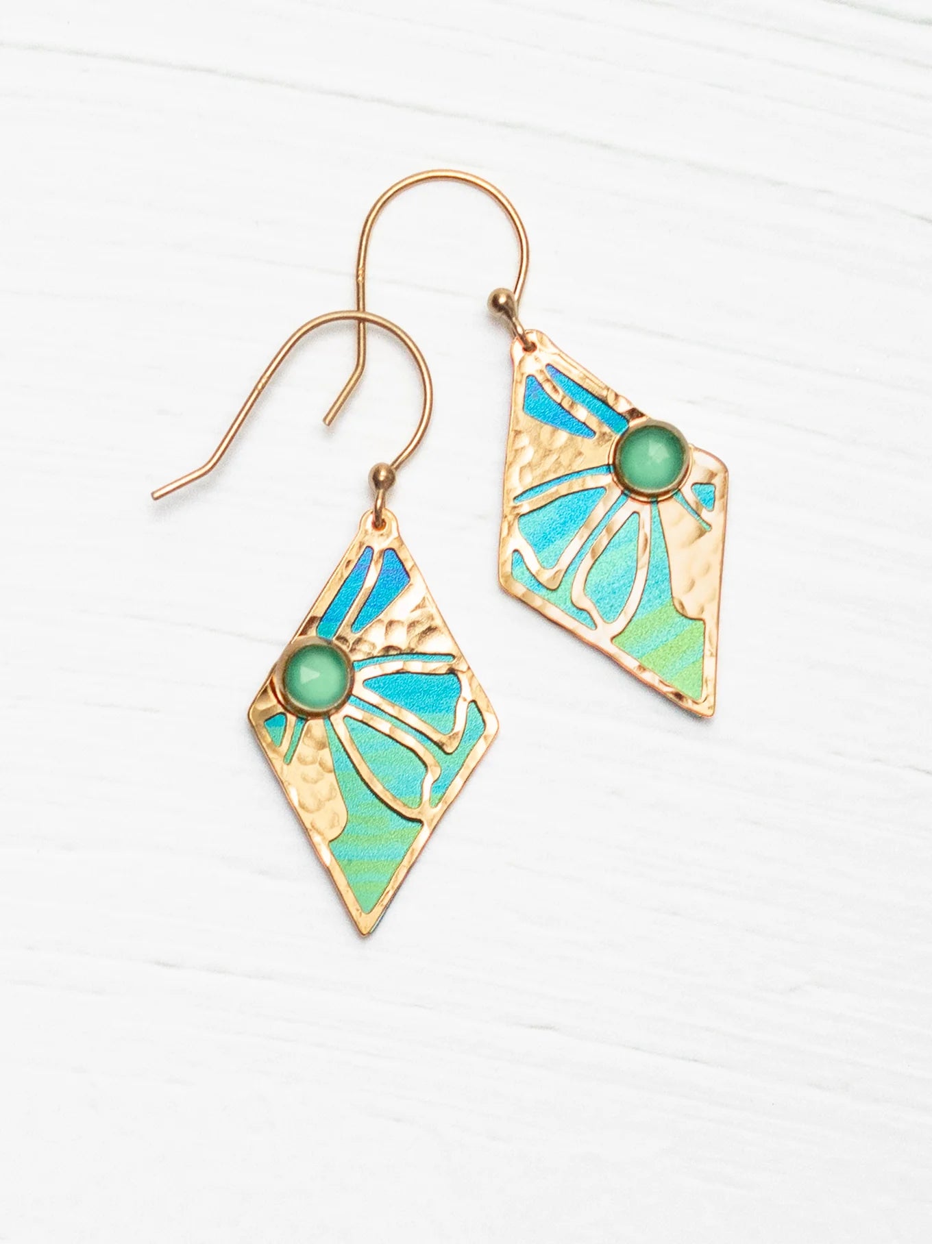 The Holly Yashi Drew Earrings are a pair of 18K gold-plated, diamond-shaped accessories featuring an intricate stained glass-like design in shades of blue and green with a central green stone. Resembling Retro floral earrings, they have delicate hooks for wearing and are beautifully displayed on a light wooden surface.