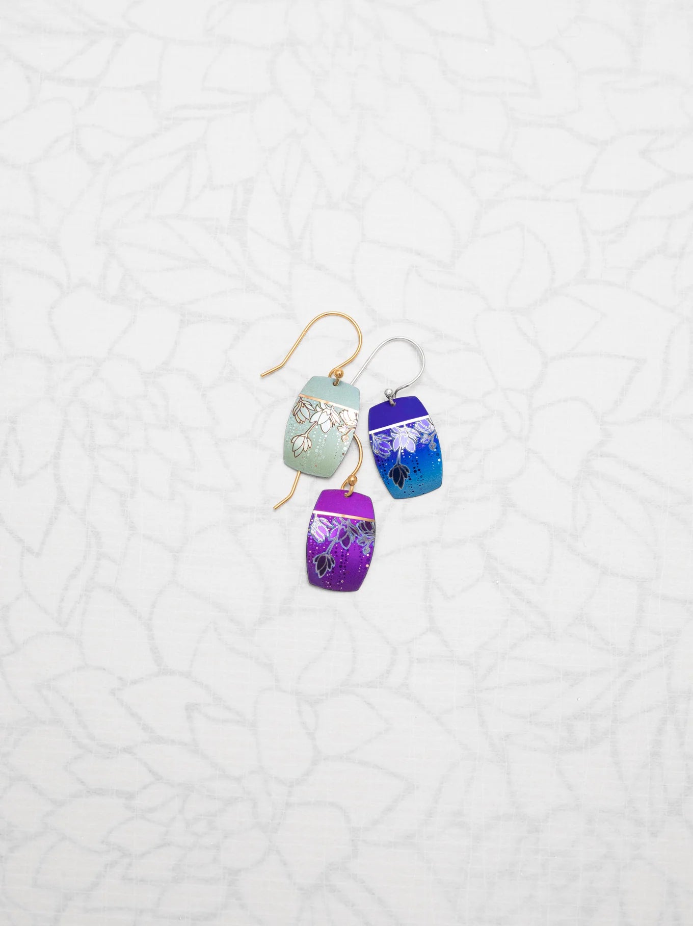 A pair of Holly Yashi Cari Earrings displayed on a white, floral-patterned background. The niobium earrings feature small, rectangular pieces with intricate designs in shades of blue, purple, and green, each attached to a gold hook.