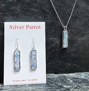 Displayed on a dark stone background are a set of sterling silver earrings and a pendant necklace, each featuring multicolored opal-like rectangular stones and crafted by Israeli silversmiths. The elegantly mounted earrings are presented on a card labeled "Silver Parrot" and "Sterling Silver." This exquisite collection is known as the Roman Glass Set In An Elegant Sterling Pendant.