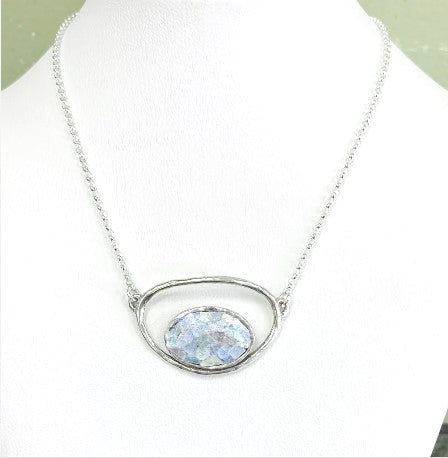 The Oval Roman Glass Sterling Silver Necklace showcases a silver chain adorned with an oval-shaped pendant featuring a textured Roman Glass gemstone at its core. The pendant is embraced by an open metal frame, offering a minimalist yet elegant aesthetic. Displayed on a white bust, this necklace evokes the timeless beauty of the desert of Israel.