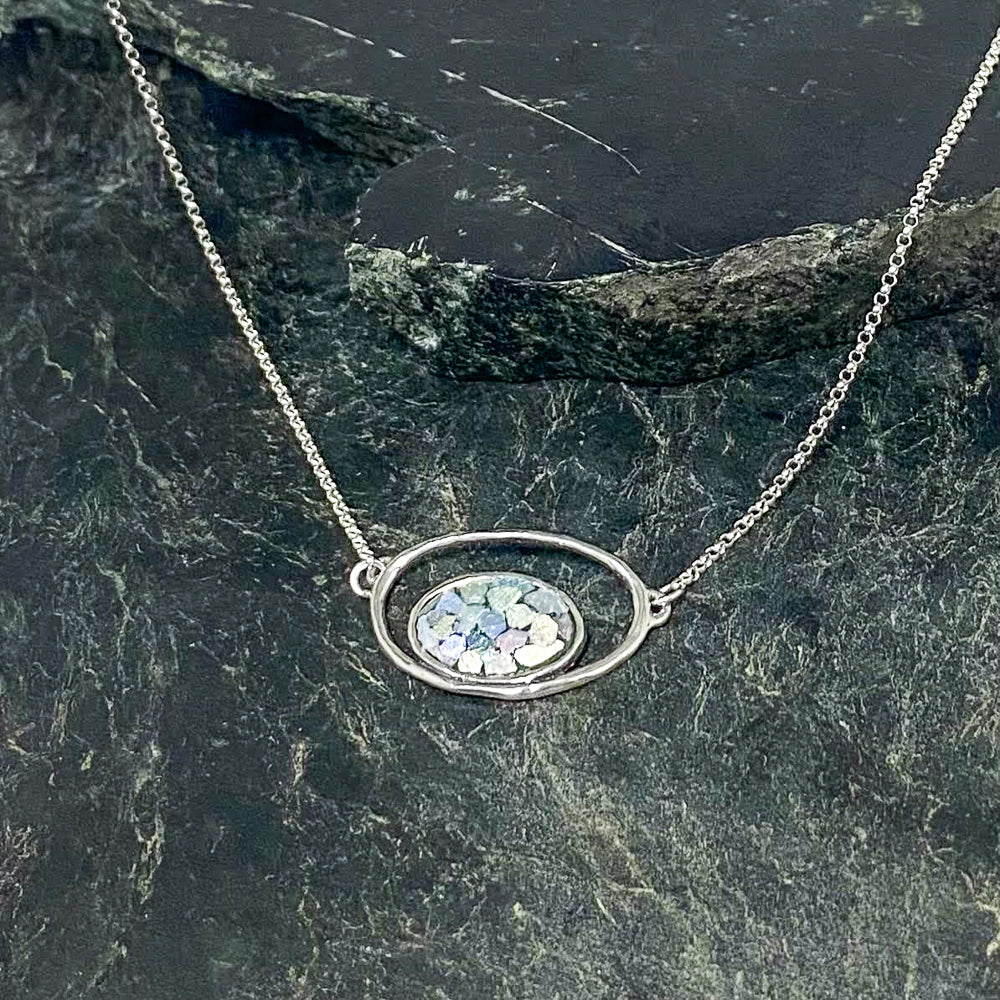 The Oval Roman Glass Sterling Silver Necklace is a delicate silver piece featuring a chain connected to a central oval pendant. The pendant showcases an intricately designed mosaic pattern with sparkling, iridescent pieces of Roman Glass set against a dark, textured background, evoking the ancient treasures from the desert of Israel.
