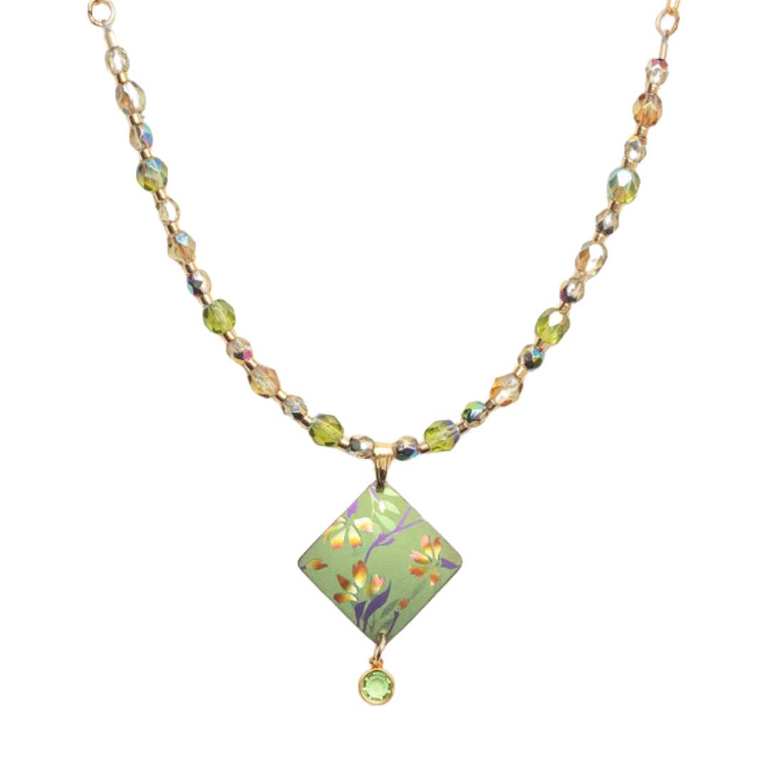 The Holly Yashi Garden Sonnet Beaded Necklace is a delicate piece featuring a chain of small, multicolored beads. At its center lies a green diamond-shaped pendant with an intricate floral design, complemented by a smaller round green gem that dangles below it. This necklace has a light and elegant appearance, perfectly capturing the essence of a serene garden.