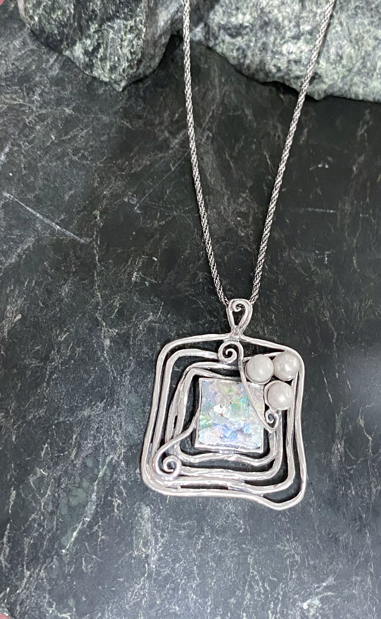 A stunning Square Roman Glass and Pearl Necklace lies on a dark marbled surface. The pendant, set in a handmade sterling setting, features an abstract, square-shaped design with intricate swirls and three small spherical accents at the top. The centerpiece is a textured, colorful Roman Glass stone.