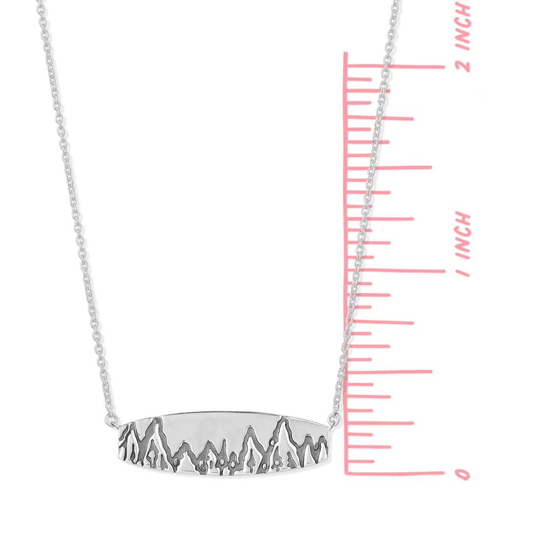 Flat rectangular sterling silver Boma necklace with engraving of mountains and trees etched along the bottom length. An adjustable rolo chain is attached to each side of the bar. Ruler is shown, seeing that pendant is one inch tall.