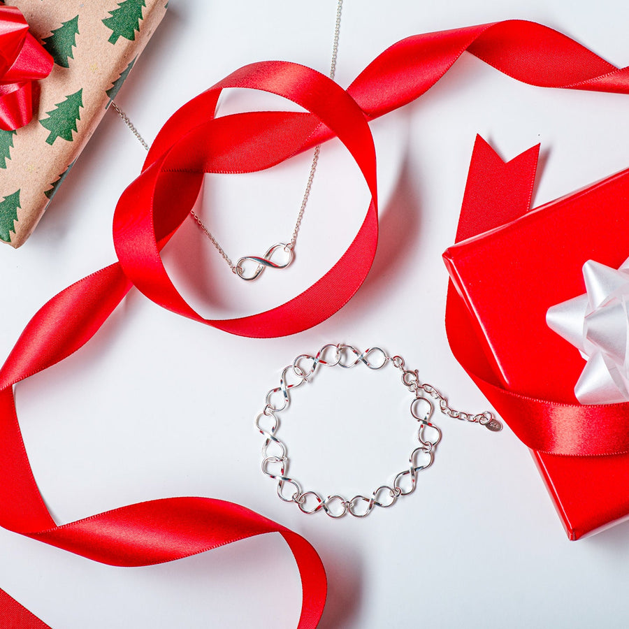 Two rock with some sticks and plants in the background, ontop of them is a pair of infinity earrings, mother and child necklace, and an ornate amethyst cuff bracelet. 