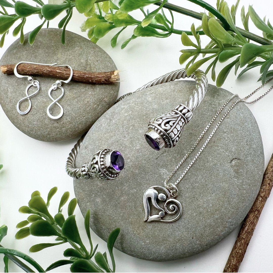 Two rock with some sticks and plants in the background, ontop of them is a pair of infinity earrings, mother and child necklace, and an ornate amethyst cuff bracelet. 