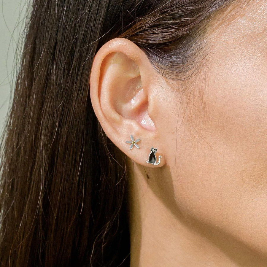 Close-up of a person's ear wearing two stud earrings from Boma Jewelry. The first earring is a small sterling silver flower, while the second earring is a Boma Black Cat Stud featuring a black and silver cat design. The person's hair is tucked behind the ear, showcasing the beautiful earrings clearly.