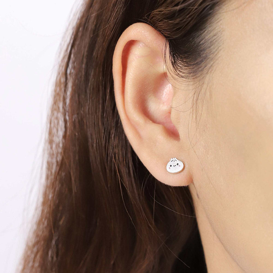 Close-up of a person wearing Boma Happy Bao Dumpling Studs, part of the sterling silver jewelry collection. The small white earring, shaped like a smiling dumpling, adds a touch of charm. The ear is shown against a plain white background, with the person's brown hair visible. Perfect foodie accessories!