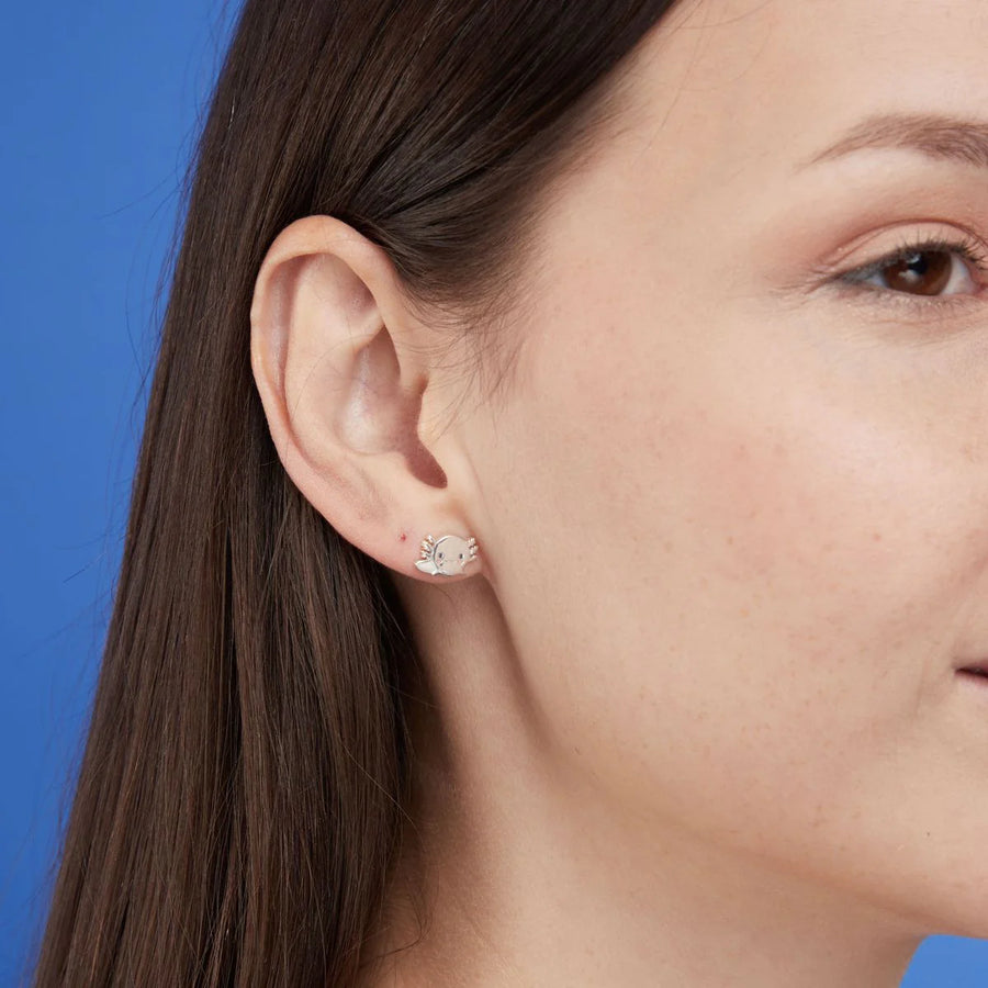 Close-up image of a person's ear adorned with Boma Cute Axolotl Studs, sterling silver earrings featuring the face of an axolotl with whiskers, a bow, and a small pink gemstone. Perfect for animal lovers and jewelry enthusiasts, the person has straight, dark brown hair and light skin against a light blue background.