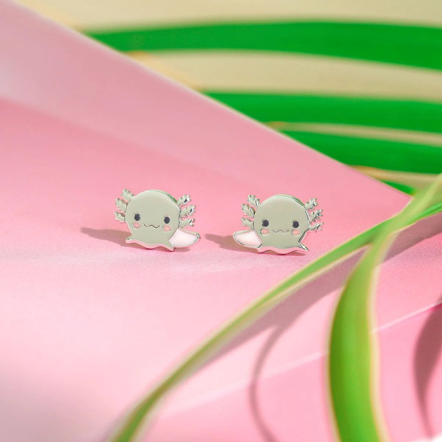 The Boma Cute Axolotl Studs, featuring adorable pink axolotls with smiling faces and pink gills, rest on a vibrant surface. Green leaves partially visible in the background add a touch of nature to the whimsical design—perfect for fans of animal-themed jewelry.