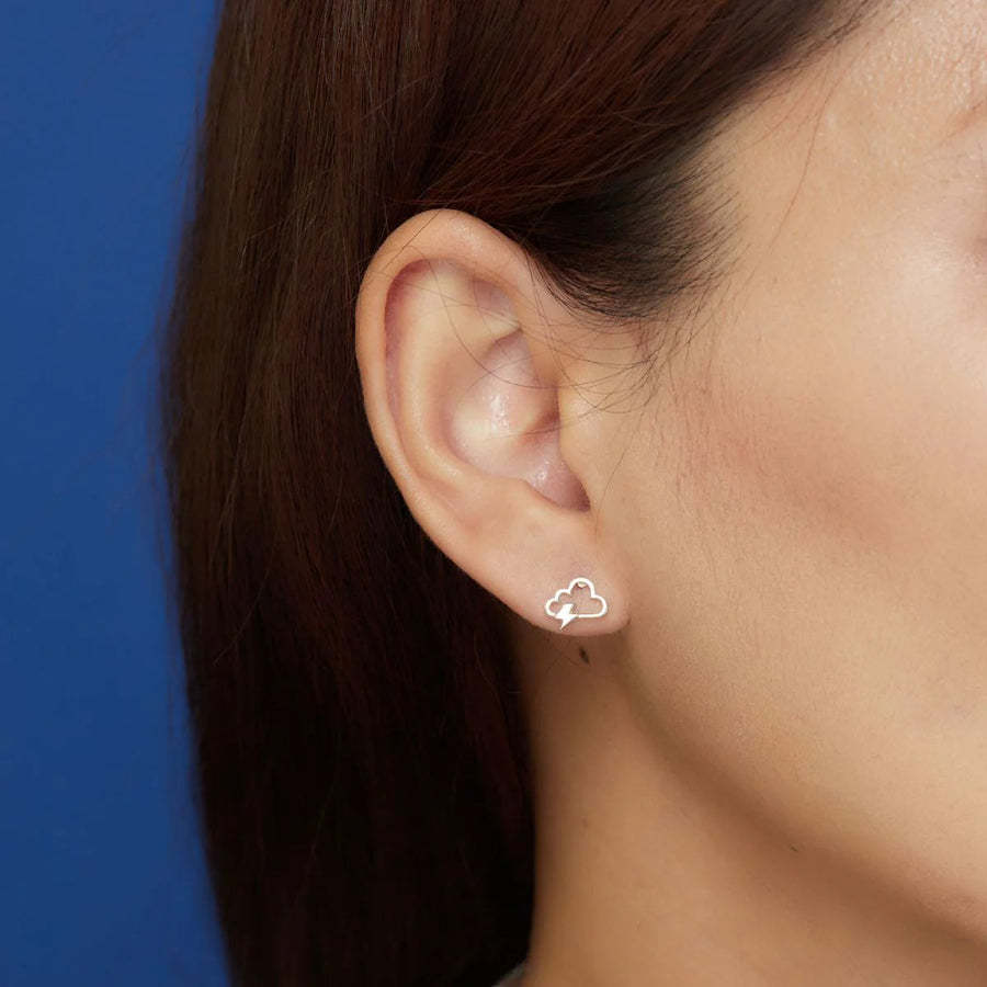 Close-up of a person's ear adorned with Boma Lightning Cloud Studs in sterling silver. The background is solid blue, and the person's dark brown hair partially covers the ear.