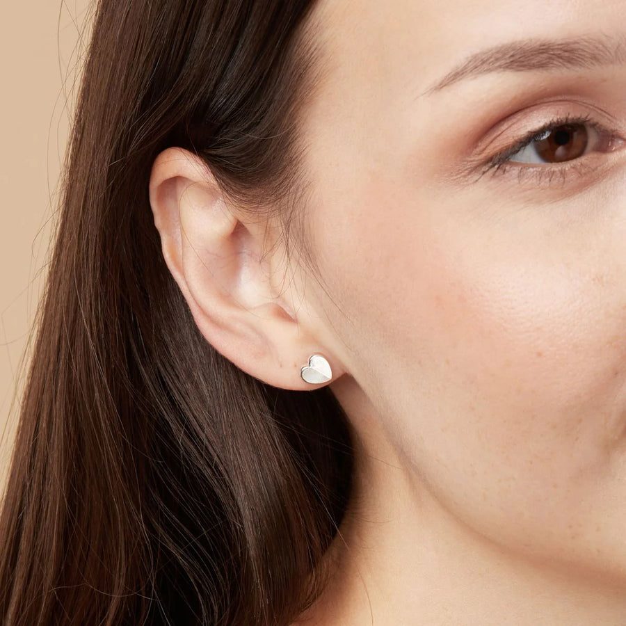 Close-up of a person wearing Boma Heart Studs with Half Stone in the left ear. The individual has straight, dark brown hair and a fair complexion. The background is a plain beige color.