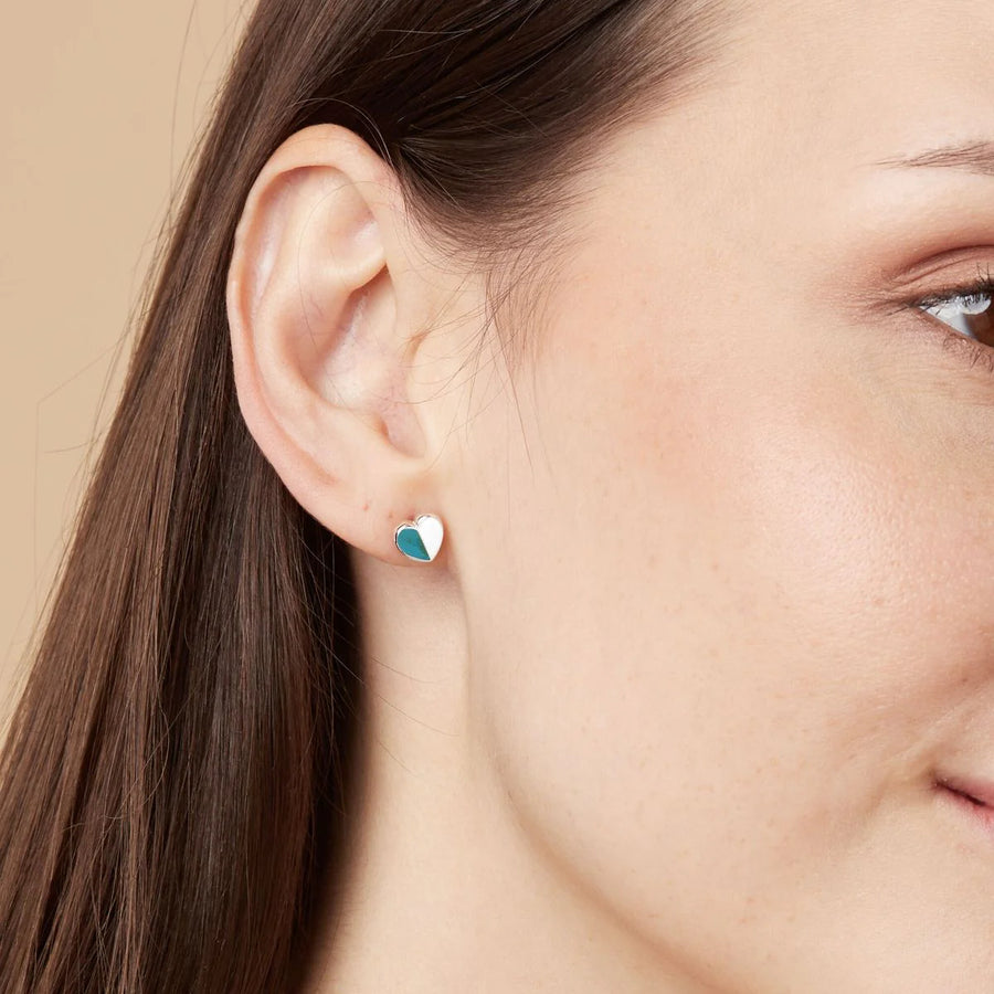 Close-up of a woman's ear wearing Boma Heart Studs with Half Stone, crafted from 925 sterling silver. The hypoallergenic earring features a small heart design split into two colors: white on the left half and teal on the right half, set against a solid light beige background.