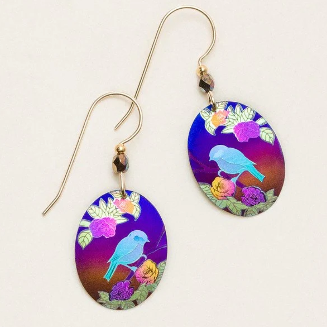 Holly Yashi Birdsong Earrings - Ovular flat Holly Yashi earring with a simple design of a bright blue-colored bird sitting on top of a purple branch with flowers and leaves. The background is a blue-to-orange gradient sky. The earrings are on a gold-filled ear wire with a bold brown bead at the top.