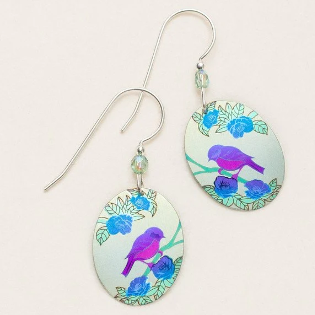 Holly Yashi Birdsong Earrings - Ovular flat Holly Yashi earring with a simple design of a purple and pink-colored bird sitting on top of a blue branch with leaves. The background is a gradient light blue sky. The earrings are on a sterling silver ear wire with a light blue bead at the top.