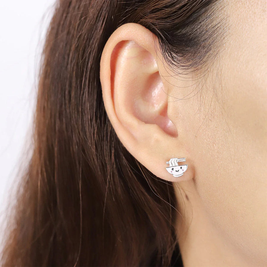 Close-up of a woman's ear adorned with quirky jewelry: the Boma Happy Ramen Studs. These sterling silver earrings, featuring an intricately designed smiling sloth hanging from a branch, stand out against a plain white wall. The detailed craftsmanship accentuates the sloth's charming facial features beautifully.