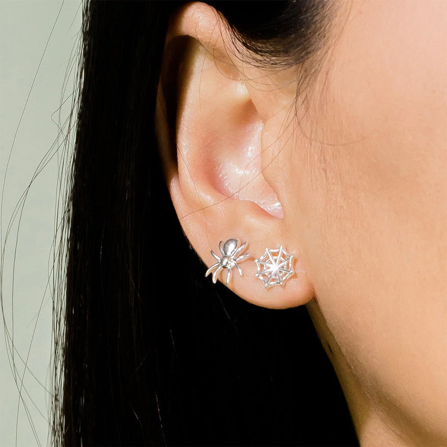 Close-up of a person's ear adorned with two Boma Spider Studs. The earring on the left is shaped like a small spider, while the one on the right resembles a spider web, both crafted from hypoallergenic sterling silver and attached to the ear lobe. The background is softly blurred.