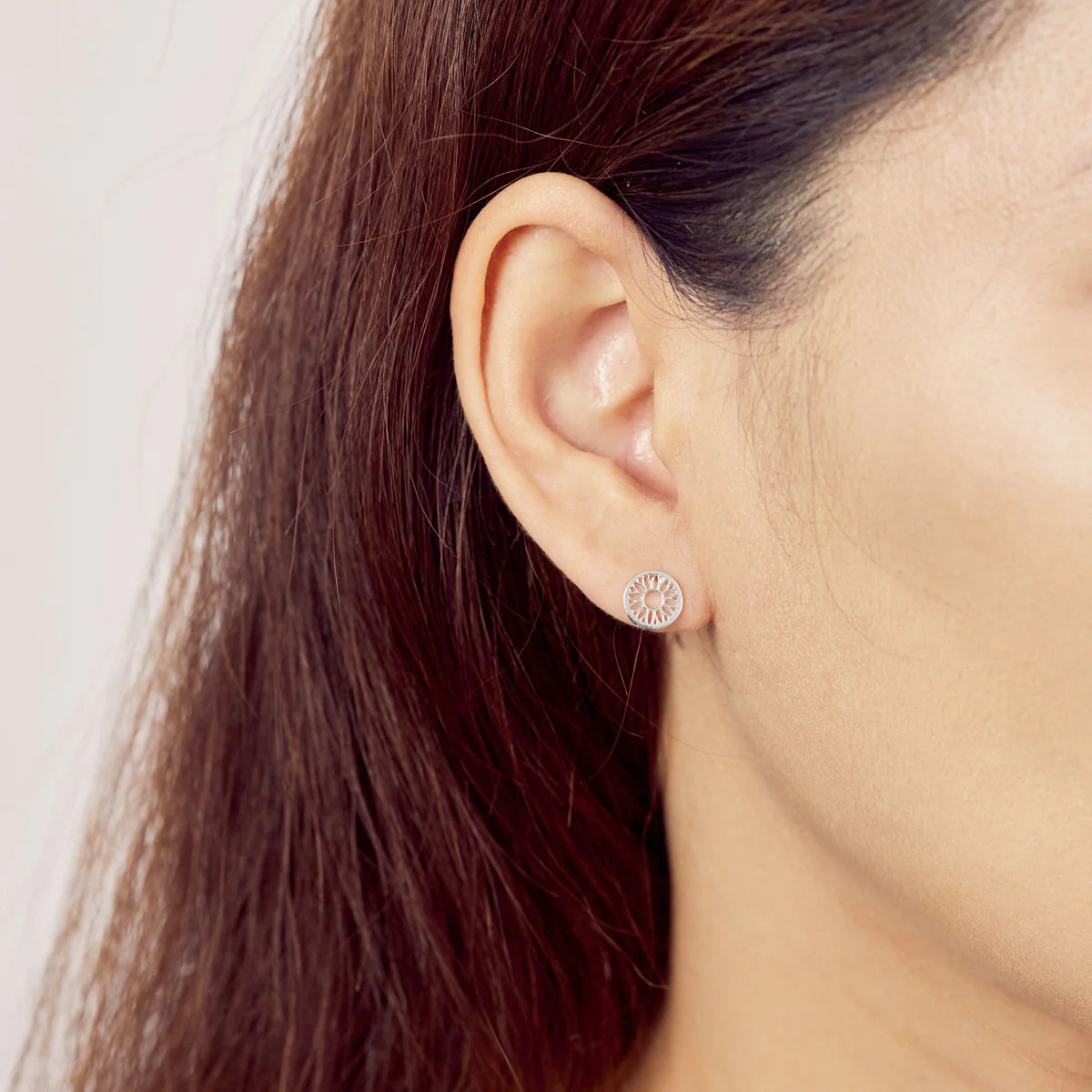 A close-up of a woman's ear showcasing the Boma Sun Flower Studs, featuring a small, round design with delicate, intricate details in 925 Sterling Silver. The woman's dark brown hair is tucked behind her ear, highlighting the hypoallergenic earrings. The background is blurred.