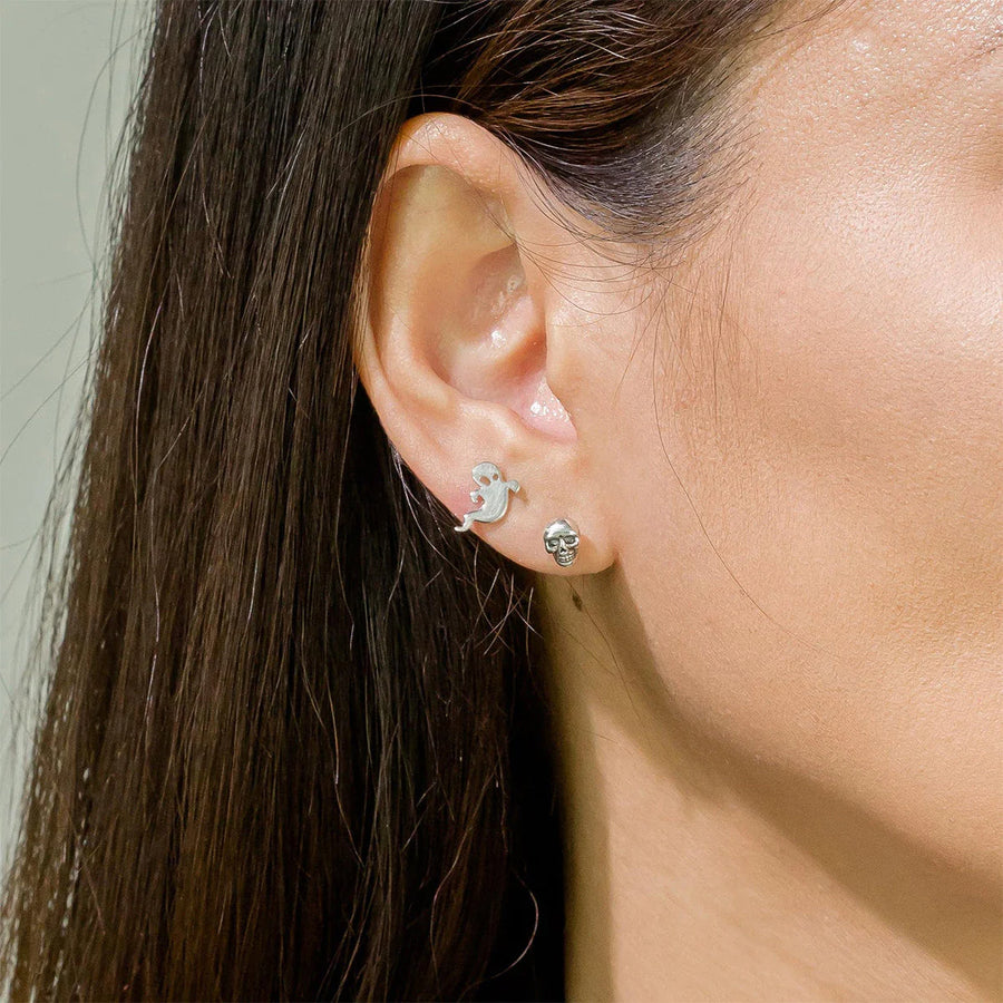 Close-up of a woman's ear adorned with two Halloween earrings. The higher earring is the Boma Spooky Ghost Stud, which is a small, sterling silver ghost-shaped stud with googly eyes, while the lower earring is a silver skull-shaped stud. Her straight brown hair partially covers her ear, adding to the spooky charm.