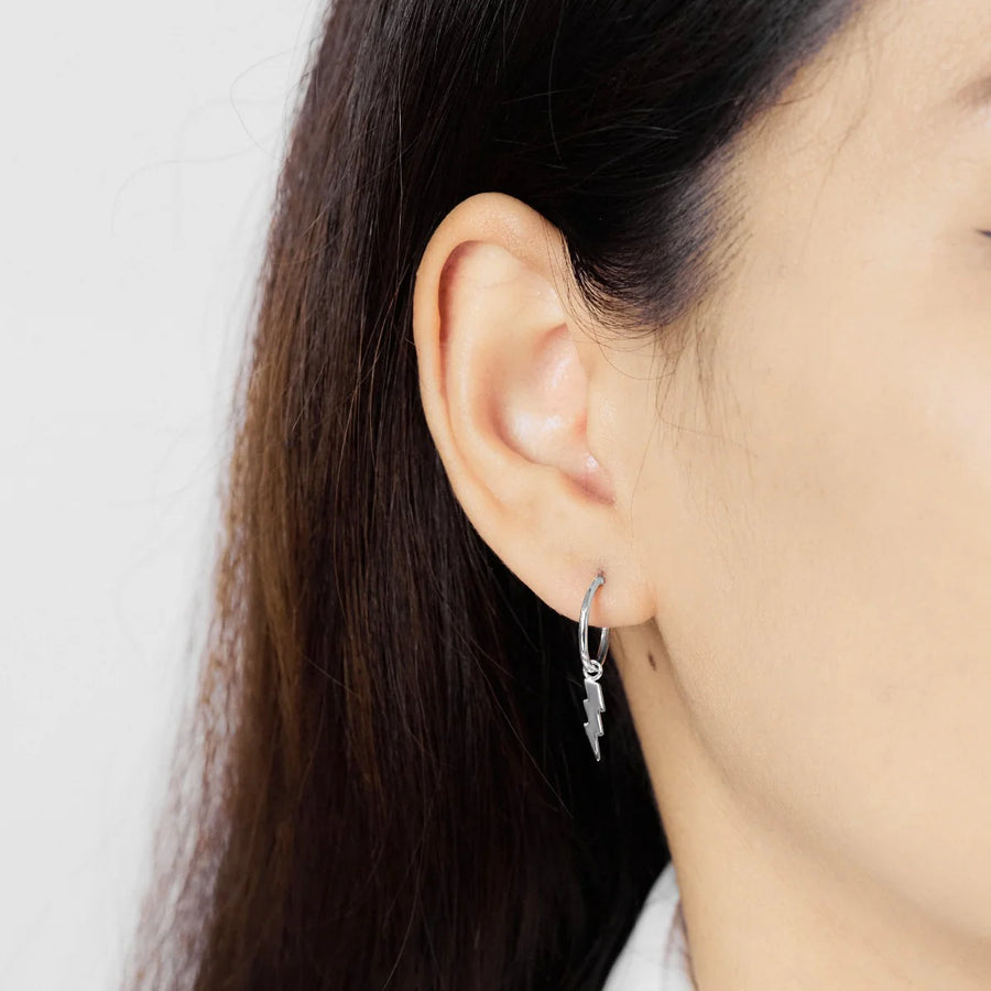 Close-up of a person with long, dark hair wearing Boma Lightning Hoops, sterling silver hoop earrings adorned with a dangling lightning bolt charm. The background is plain and light-colored. These hypoallergenic earrings ensure comfort while showcasing unique lightning bolt designs.