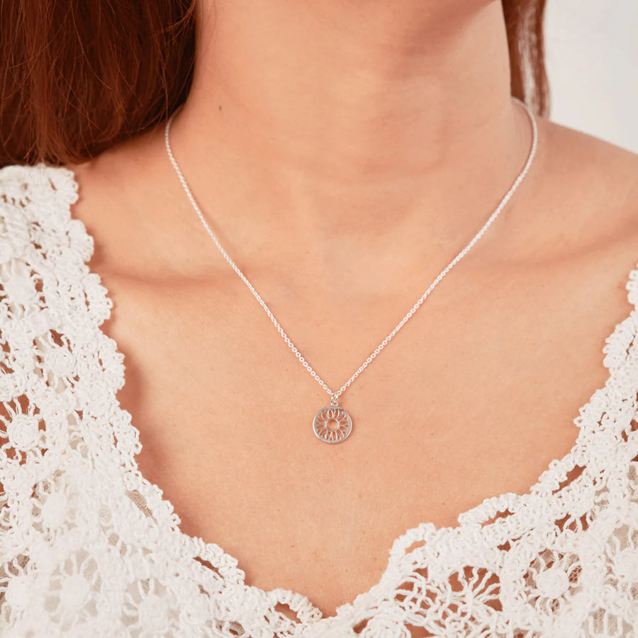 A person wearing a delicate Boma Sun Flower Necklace made of 925 sterling silver with a circular pendant featuring intricate designs. The hypoallergenic piece complements their white, lace-patterned top beautifully.