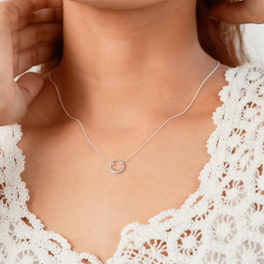 A close-up of a person wearing the elegant Boma Happy Face Necklace, delicately crafted from 925 Sterling Silver. Dressed in a white, lace-textured top, their hands are raised, touching the sides of their neck. The background is softly blurred, complementing the elegance of the matching earrings they wear.