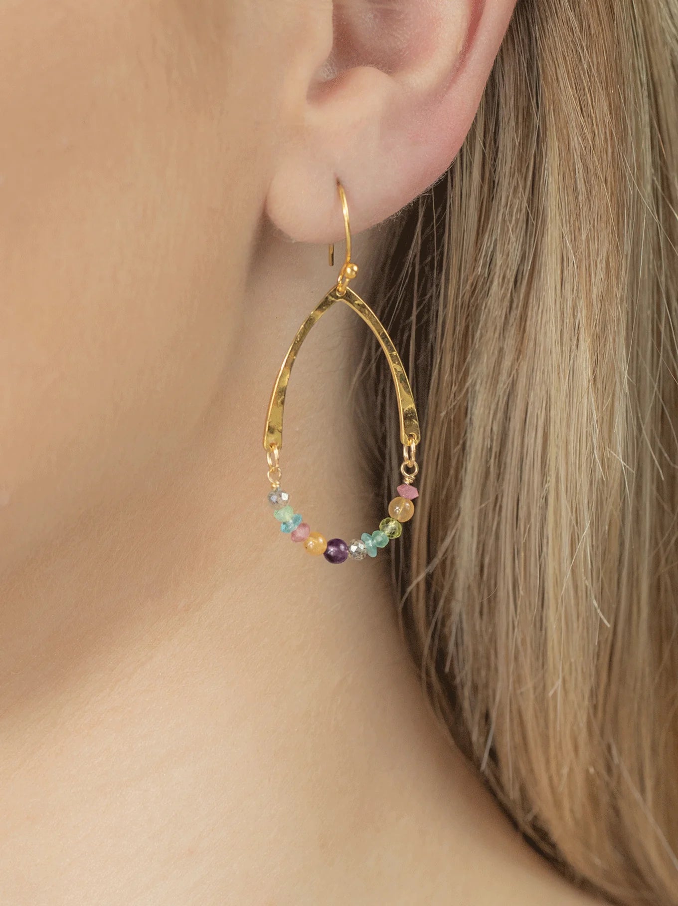 Close-up of a person's ear wearing a gold hoop earring in a teardrop shape. The Holly Yashi Mikayla Dangles are 18K Gold-plated and adorned with small, colorful beads arranged in a scalloped pattern along the lower section of the hoop. The person's hair is light-colored and pulled back.