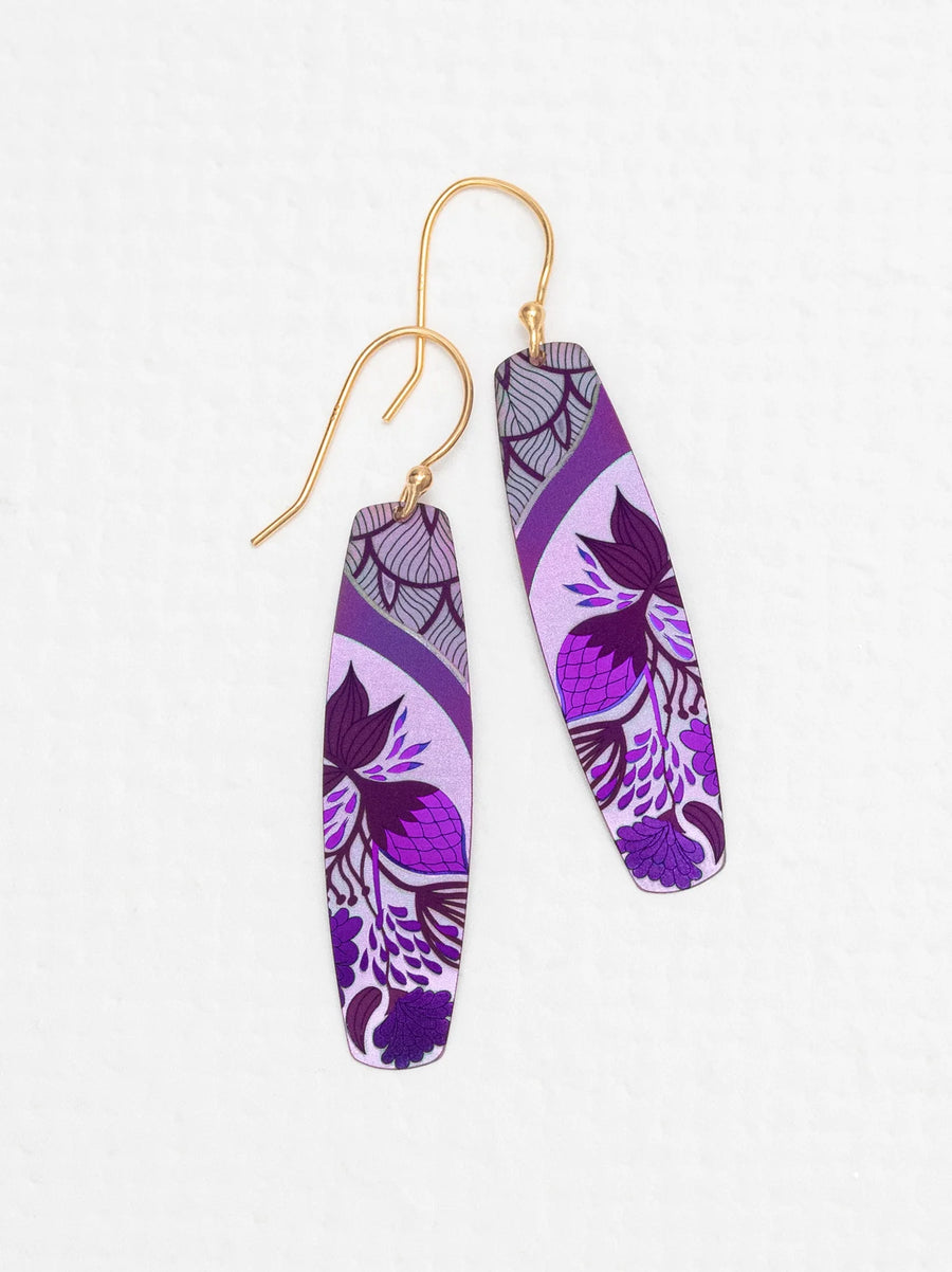 A pair of Holly Yashi Malia Earrings featuring elongated oval pendants with vibrant purple tropical florals on a lilac background, accented with intricate patterns and 18K Gold-plated Niobium hooks. The earrings lay flat on a white surface.