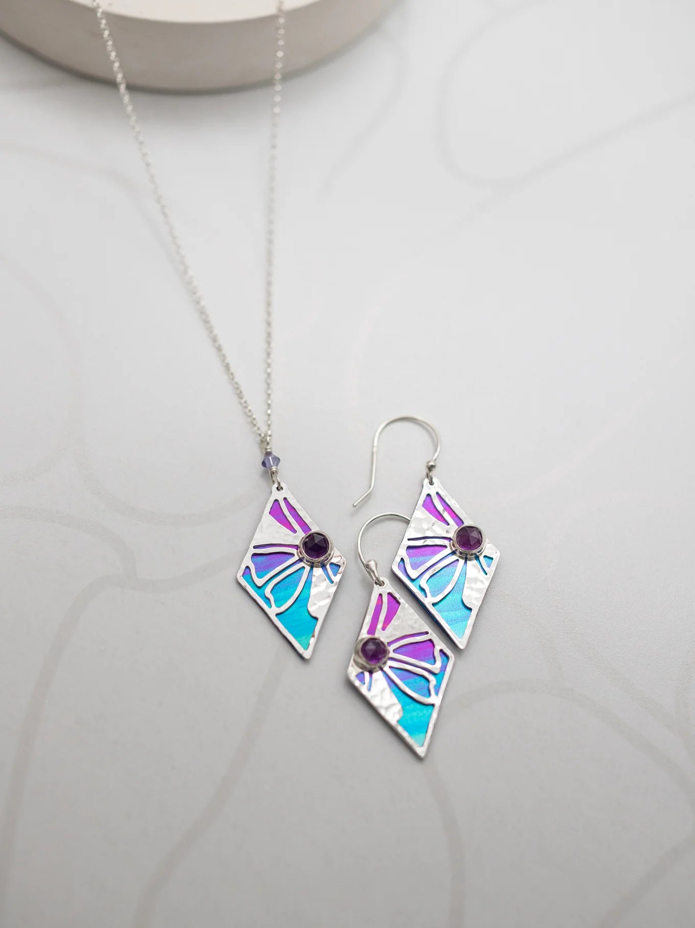 The Holly Yashi Drew Necklace and matching earrings set boasts a geometric design with segments in purple, blue, and silver. Each piece is adorned with a small central purple gemstone and crafted from gold-plated niobium against a light gray background featuring a subtle pattern.