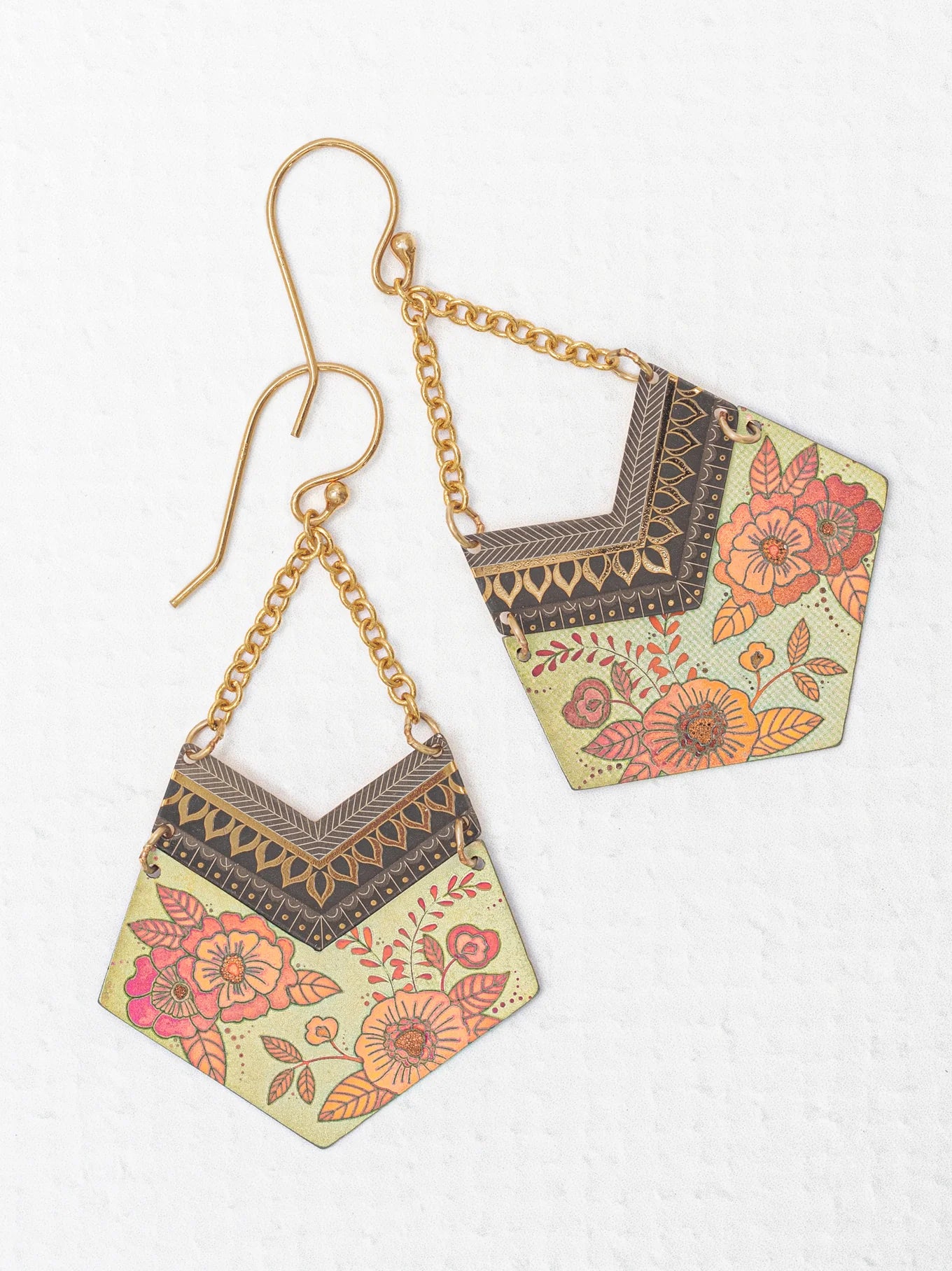 A pair of Holly Yashi Avant Garden Earrings showcasing pentagon-shaped pendants adorned with intricate floral patterns in vibrant hues of orange and pink. These boho-chic statement earrings are attached to 18K gold-plated hooks and chains, set against a textured white background.