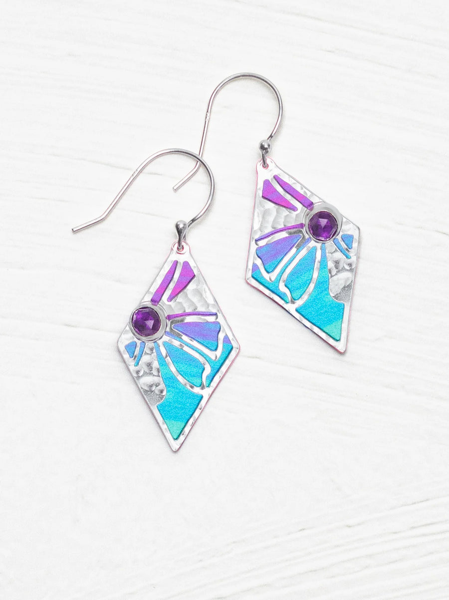 A pair of Holly Yashi Drew Earrings, 18K gold-plated dangle style with a diamond-shaped design. Each earring showcases purple and turquoise enamel inlays, a small purple gemstone, and silver accents on all visible surfaces. The background is a white textured surface.