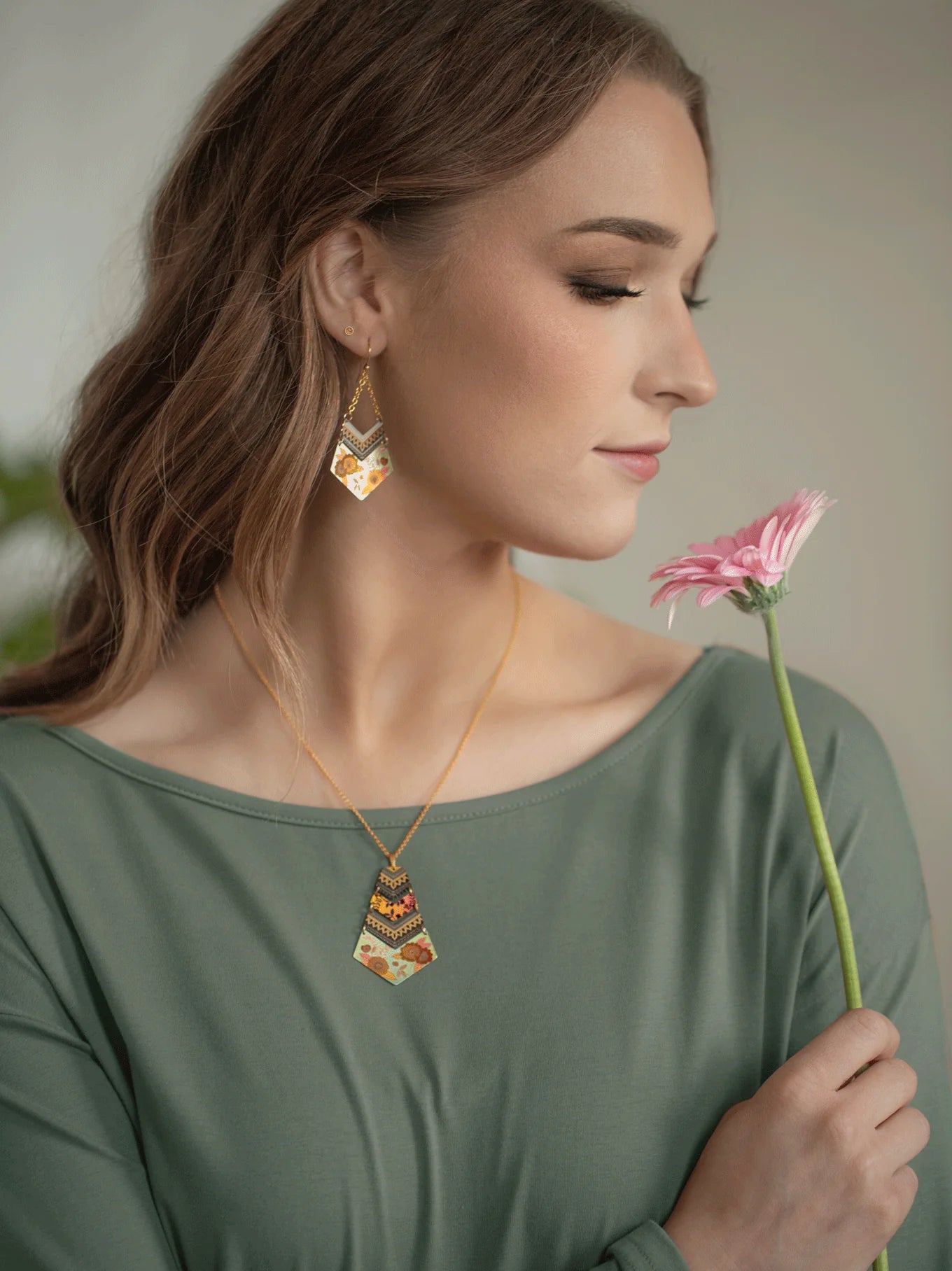 A woman with long brown hair, clad in a green top and boho-chic statement jewelry, delicately holds a pink flower near her face. She is wearing matching Holly Yashi Avant Garden Earrings and a necklace adorned with an intricate, colorful pattern. Her eyes are closed, exuding tranquility.