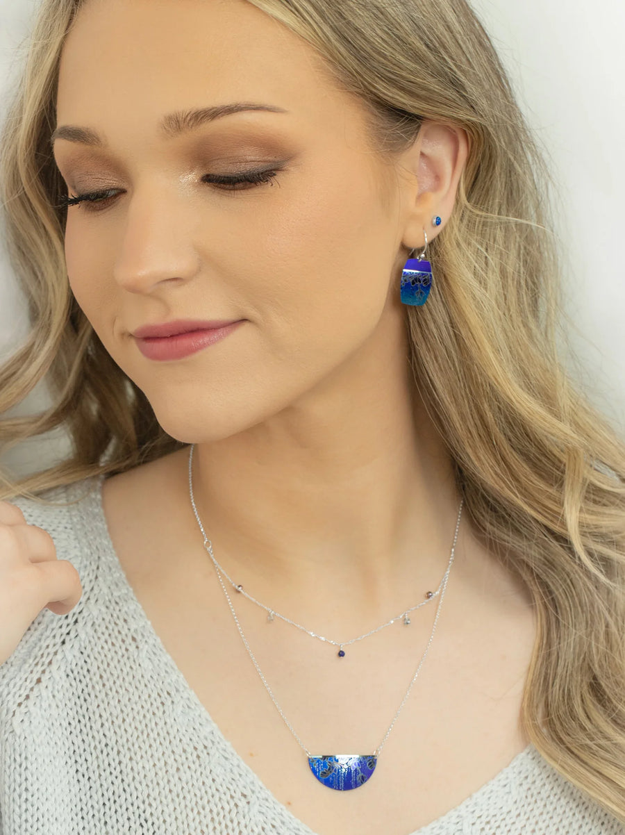 A woman with long blonde hair gently smiles with her eyes closed. She is wearing blue and silver drop earrings and a matching Holly Yashi Magnolia Necklace with blue accents. Dressed in a light-colored, textured blouse, she exudes elegance against the plain, light background.