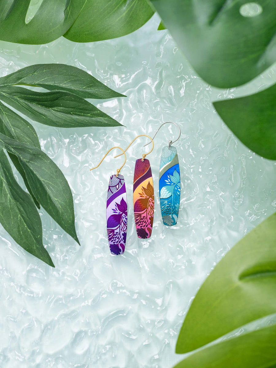 Displayed against a textured background, the Holly Yashi Malia Earrings showcase an artistic tropical floral design in vibrant colors. Surrounding the earrings are lush green leaves, and they feature delicate 18K Gold-plated Niobium hooks for wearing.