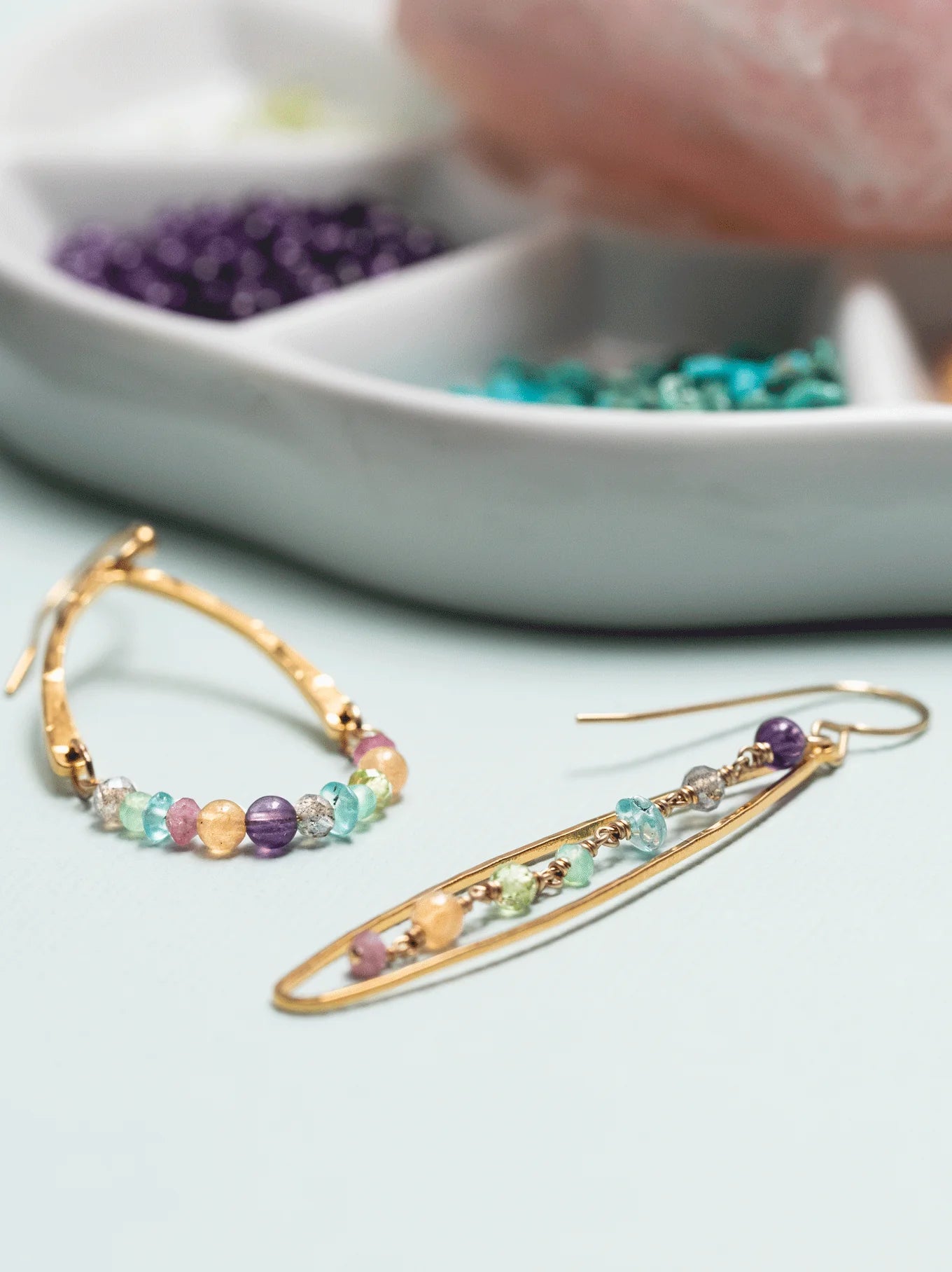 A close-up of the Holly Yashi Mikayla Dangles, showcasing multicolored beads and 18K gold-plated accents. One earring features a teardrop shape while the other displays an elongated oval shape. In the background, a dish with compartments holds purple, turquoise, and green beads.