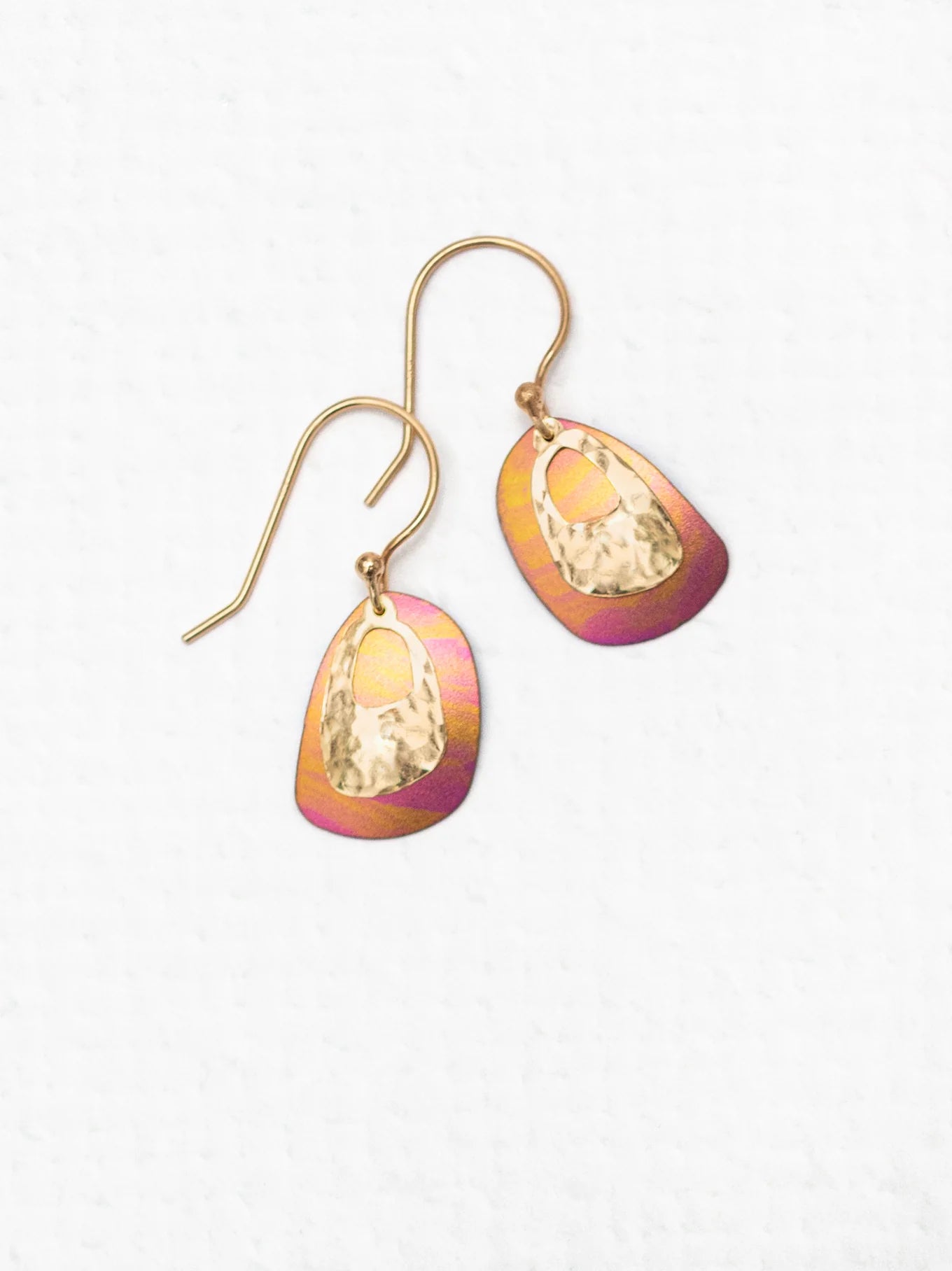 The Holly Yashi Blake Earrings are a stunning pair of dangle earrings featuring gold hooks and oval pendants with a beautiful blend of gold and magenta hues. The textured surface of the pendants is enhanced by an inner teardrop-shaped cutout, creating a captivating layered effect. Perfect for those who appreciate vibrant style, these earrings add flair to any outfit.